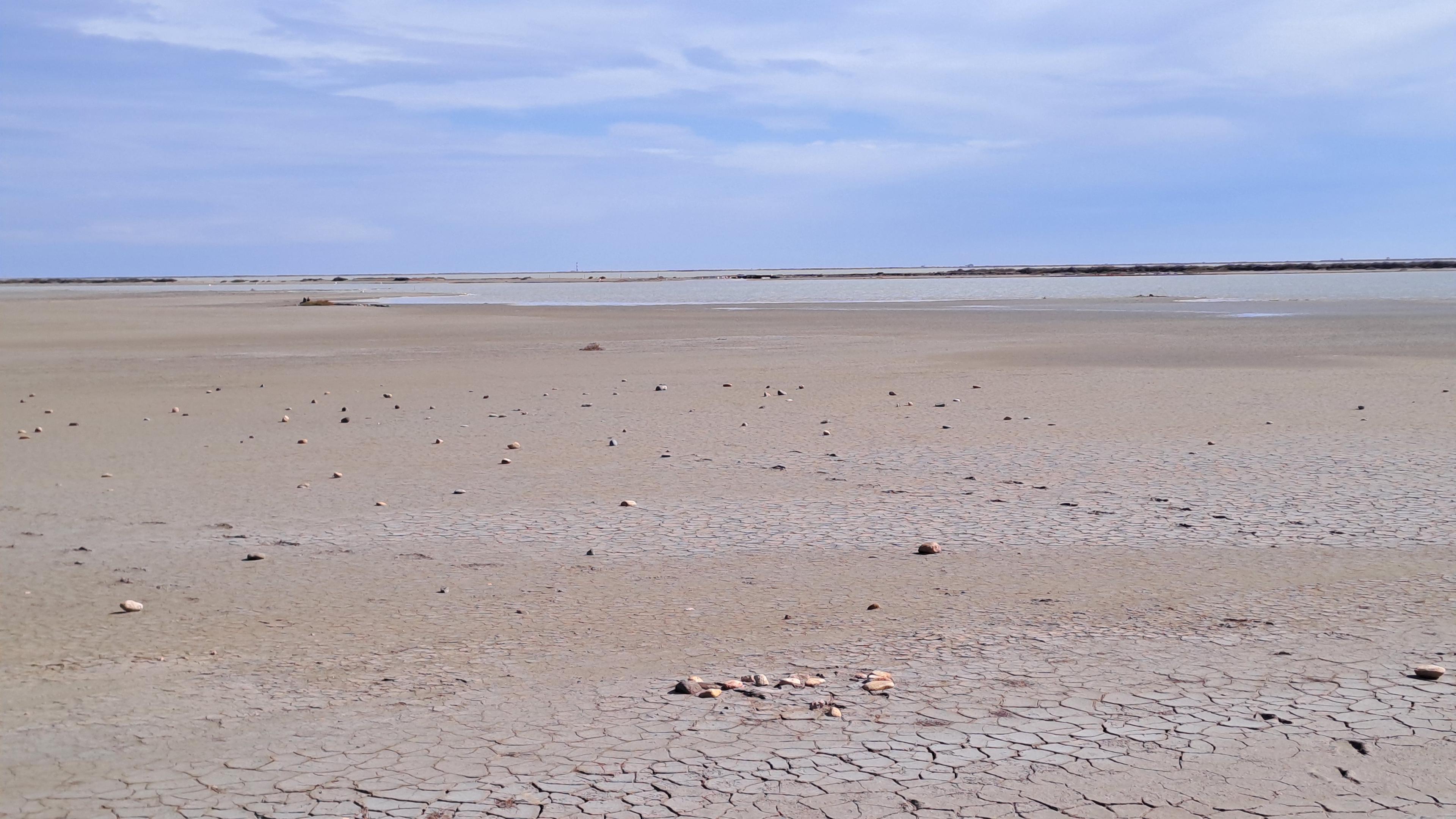 Das Bild zeigt einen fast endlos erscheinenden Sandboden und einen bewölkten Himmel in zartem Hellblau. Im Vordergrund ist der Boden durch Austrocknung eines Sees aufgebrochen und es liegen kleinere Steine am Boden.