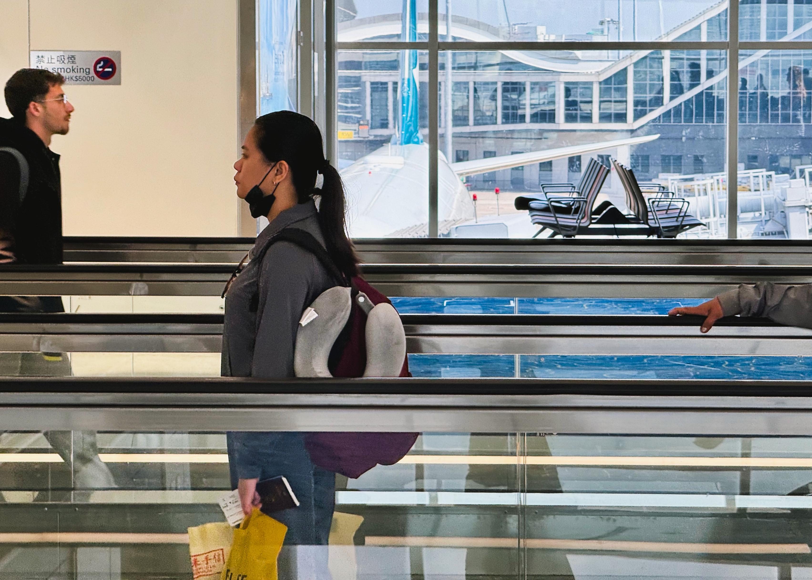 Two travelers standing on an airport moving walkway, with luggage and facing each other. Large windows reveal an airplane and airport exterior.