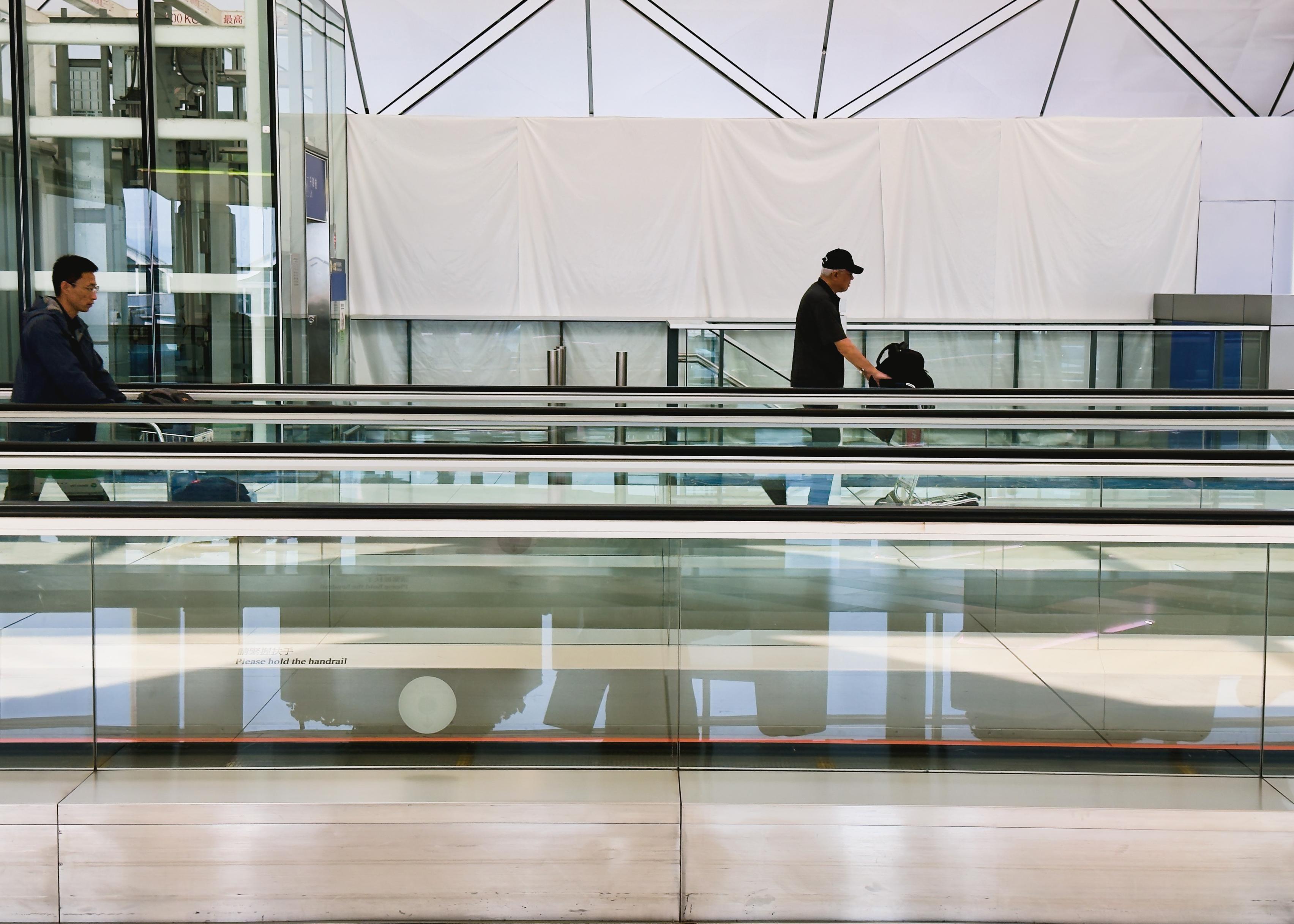 Two men with suitcases in an airport terminal, walking on separate escalators with glass sides and a modern, angular design overhead.