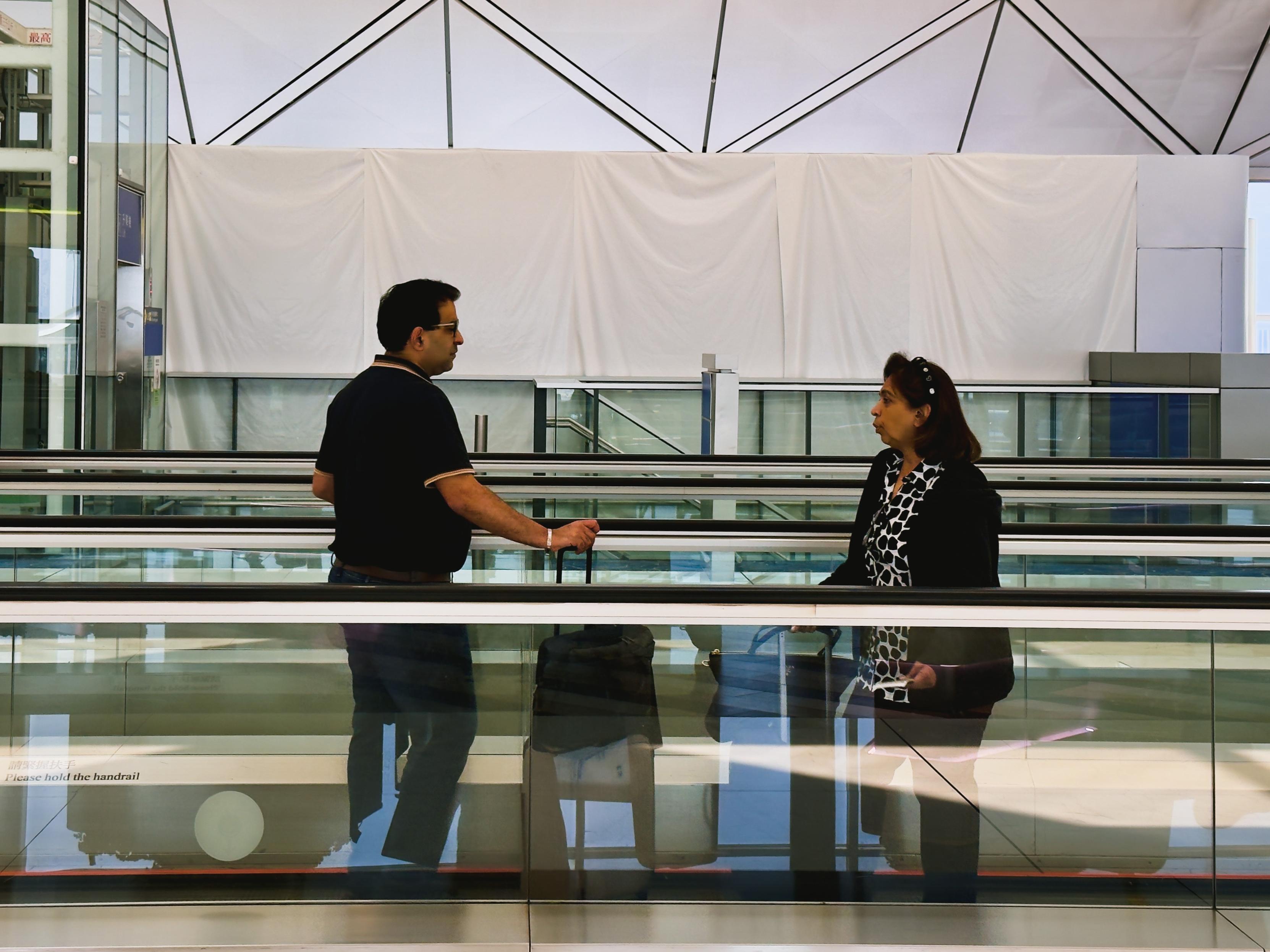 Two people standing on a moving walkway at an airport, engaged in conversation.