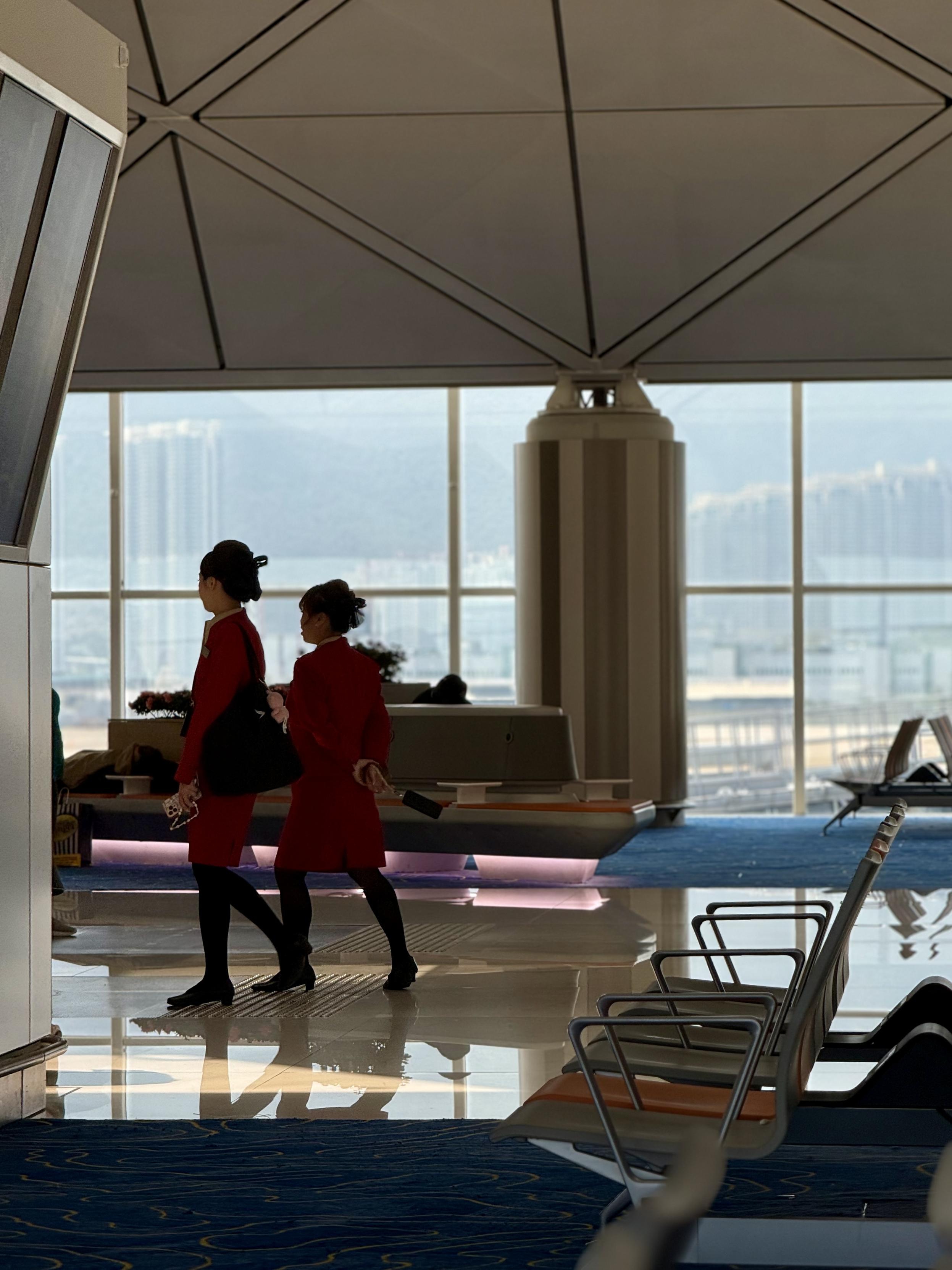 两名空乘人员穿着红色制服走过机场候机厅，背景是大窗户和城市景观。