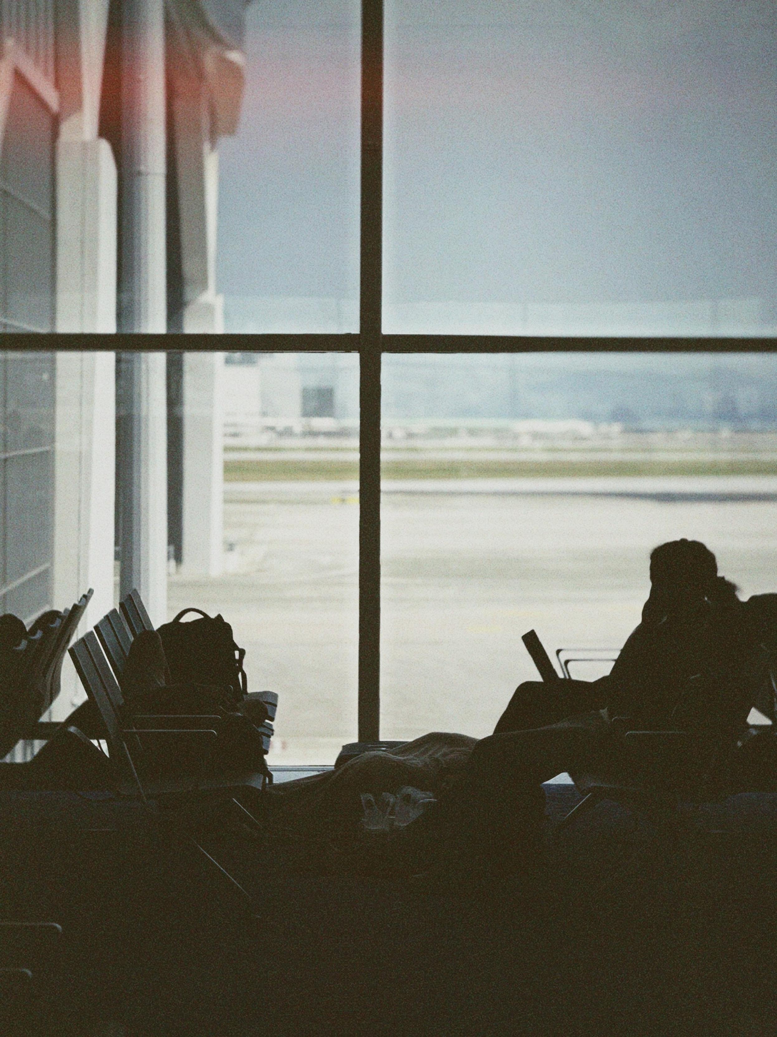 A silhouette of a person with a laptop sitting in an airport terminal, with large windows overlooking the runway.