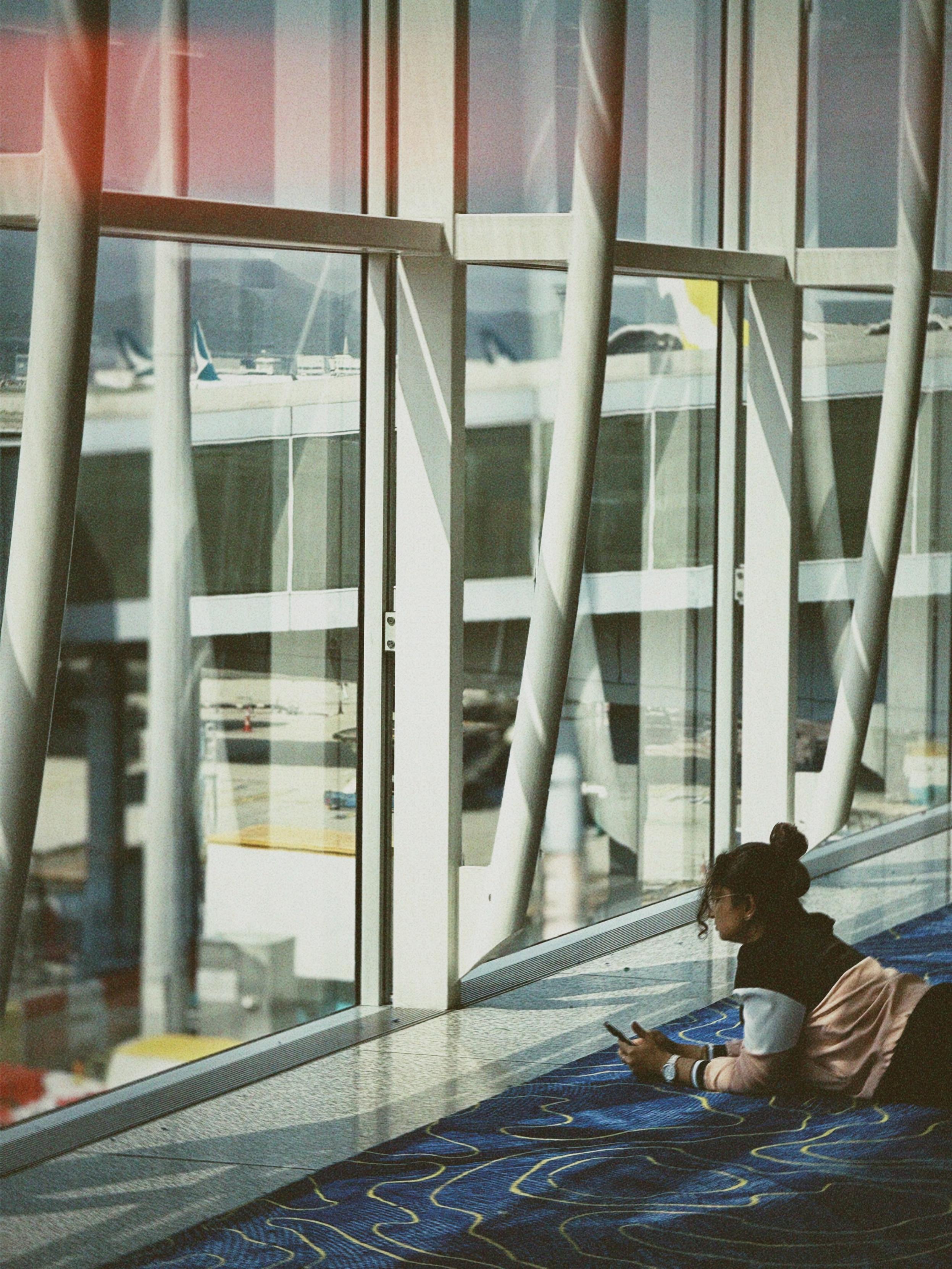 A person lies on the airport floor using a phone, near large glass windows with structural beams and planes visible outside.