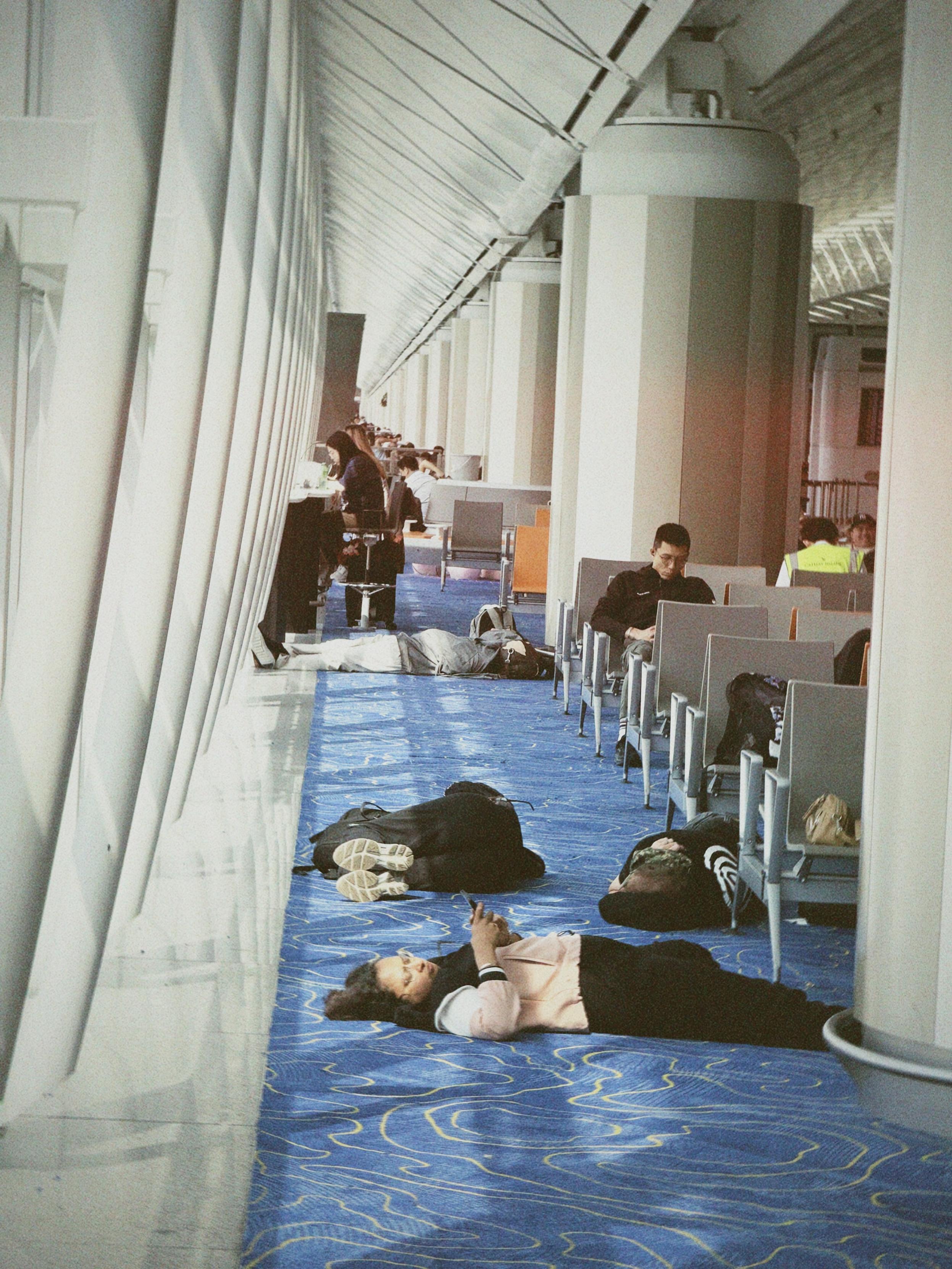 People lying and sleeping on the blue carpeted floor of an airport terminal, with others seated at counters and working on laptops.
