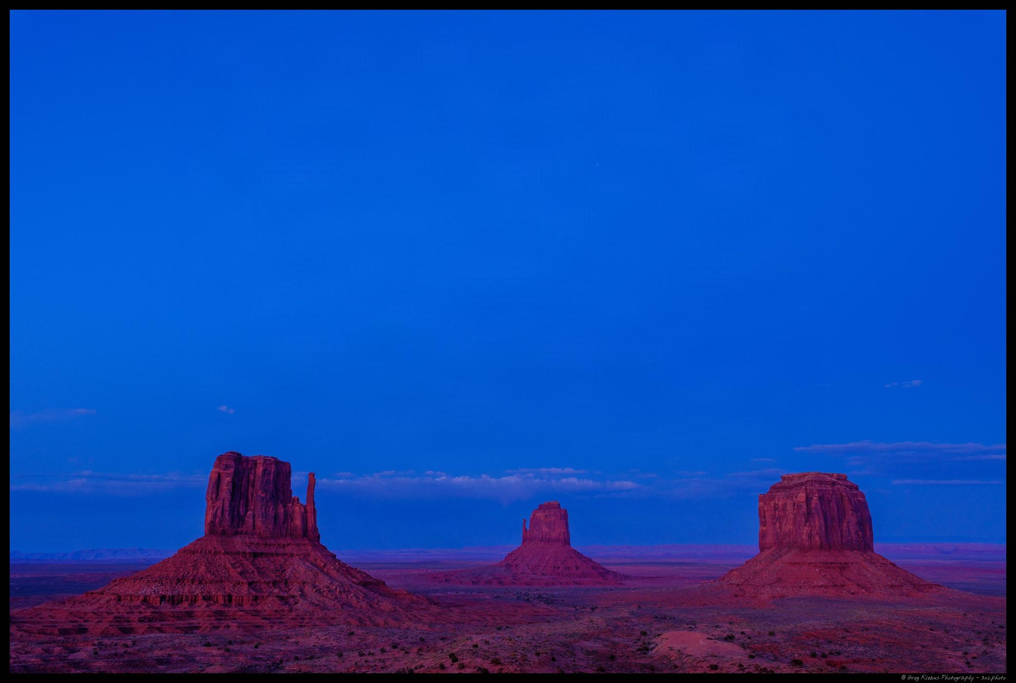 Three red sandstone buttes in the Monument Valley rise from a vast desert landscape beneath a deep blue sky.