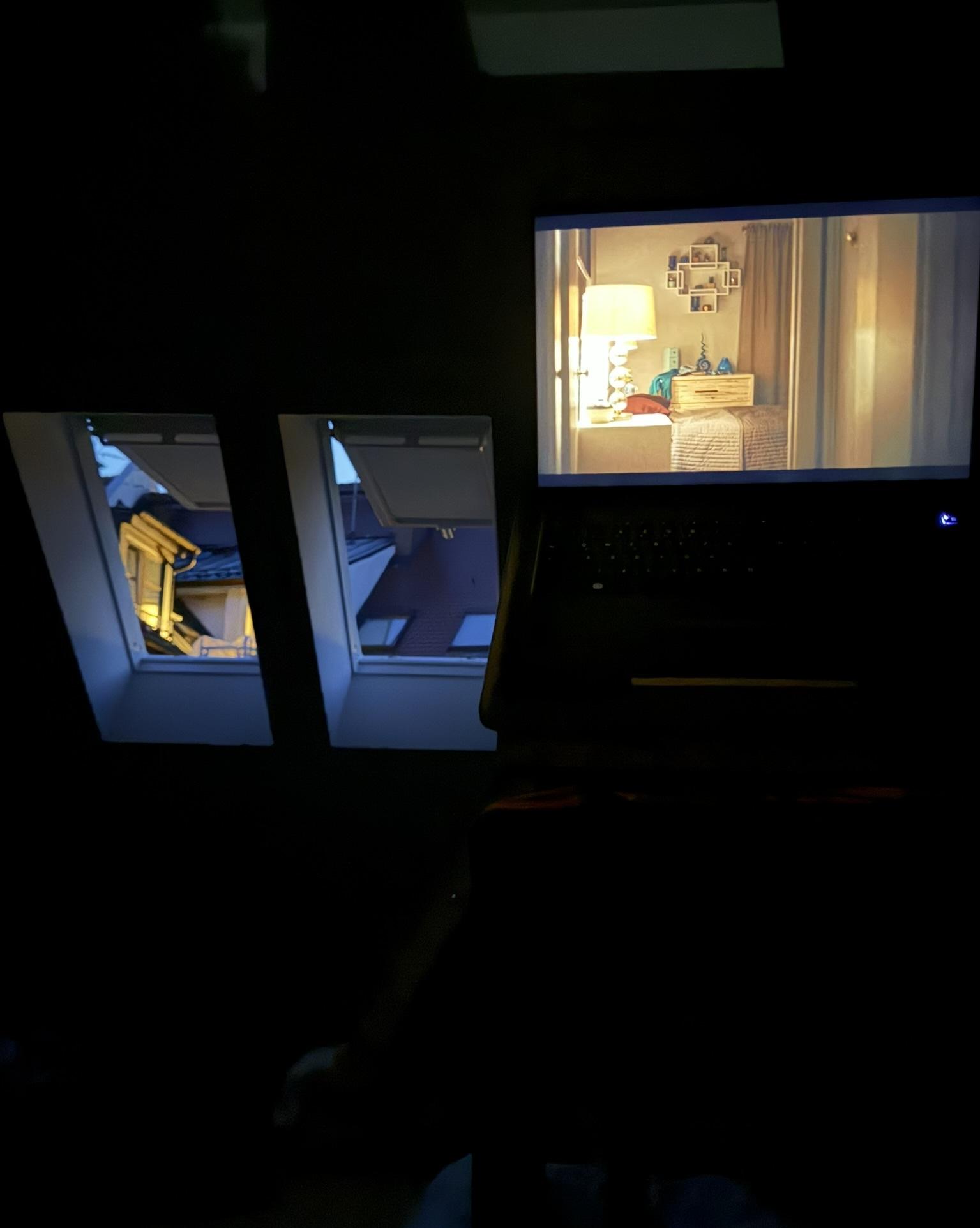 A dark room with two open roof windows on the left and a glowing computer screen on the right. The windows reveal neighboring rooftops and evening light; the screen shows a warmly lit bedroom.