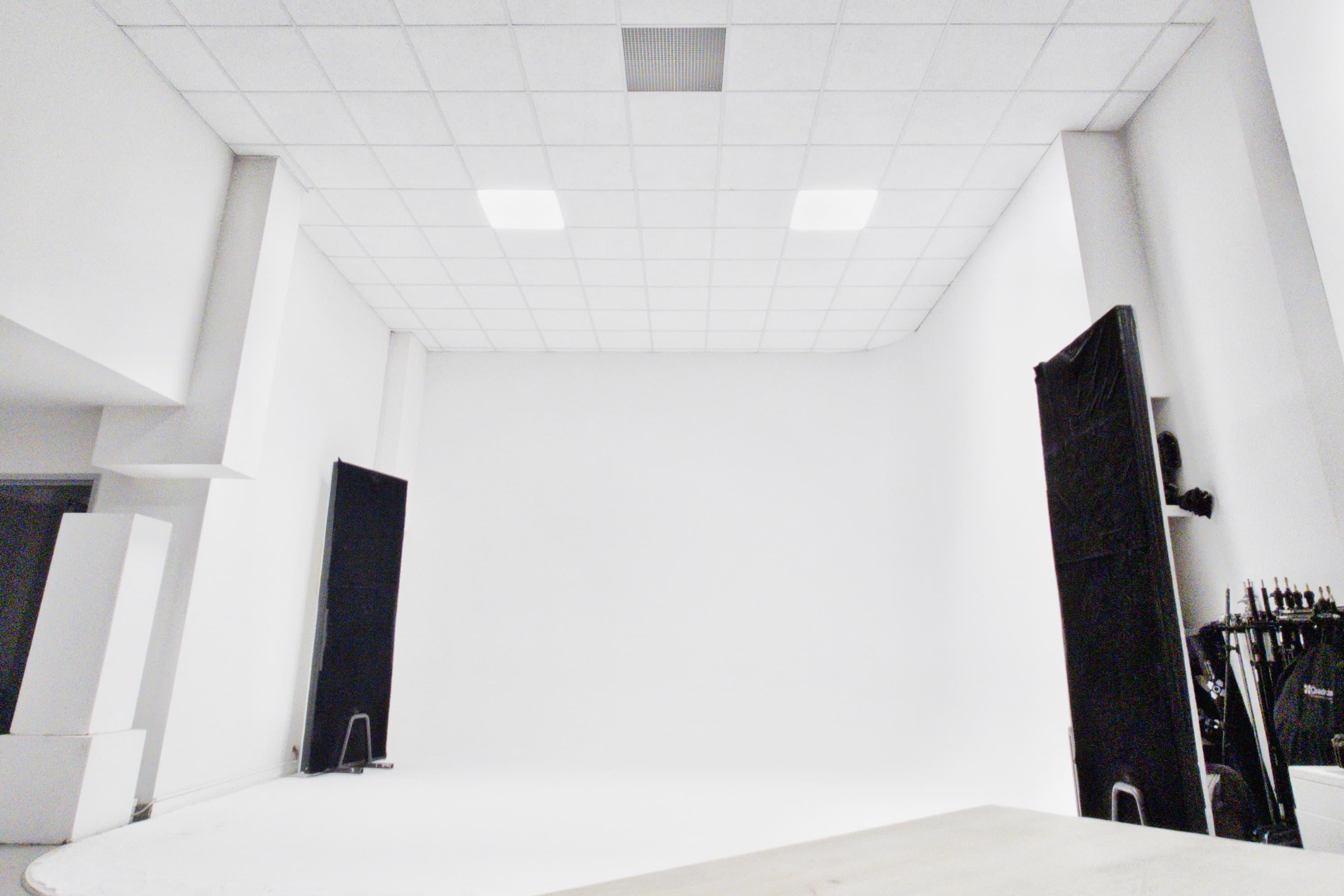 Kadr przedstawia wnętrze studia fotograficznego - Studio Stawki, z białą cykloramą - płynnym przejściem ściany w podłogę. Sufit podwieszany wyposażony jest w dwa kwadratowe panele oświetleniowe LED. Na obrzeżach cykloramy ustawione są dwie wysokie, czarne zastawki oraz zestaw statywów i sprzętu studio po prawej stronie. Wszystkie płaszczyzny są czyste, a studio pozostaje puste i gotowe do planu zdjęciowego.