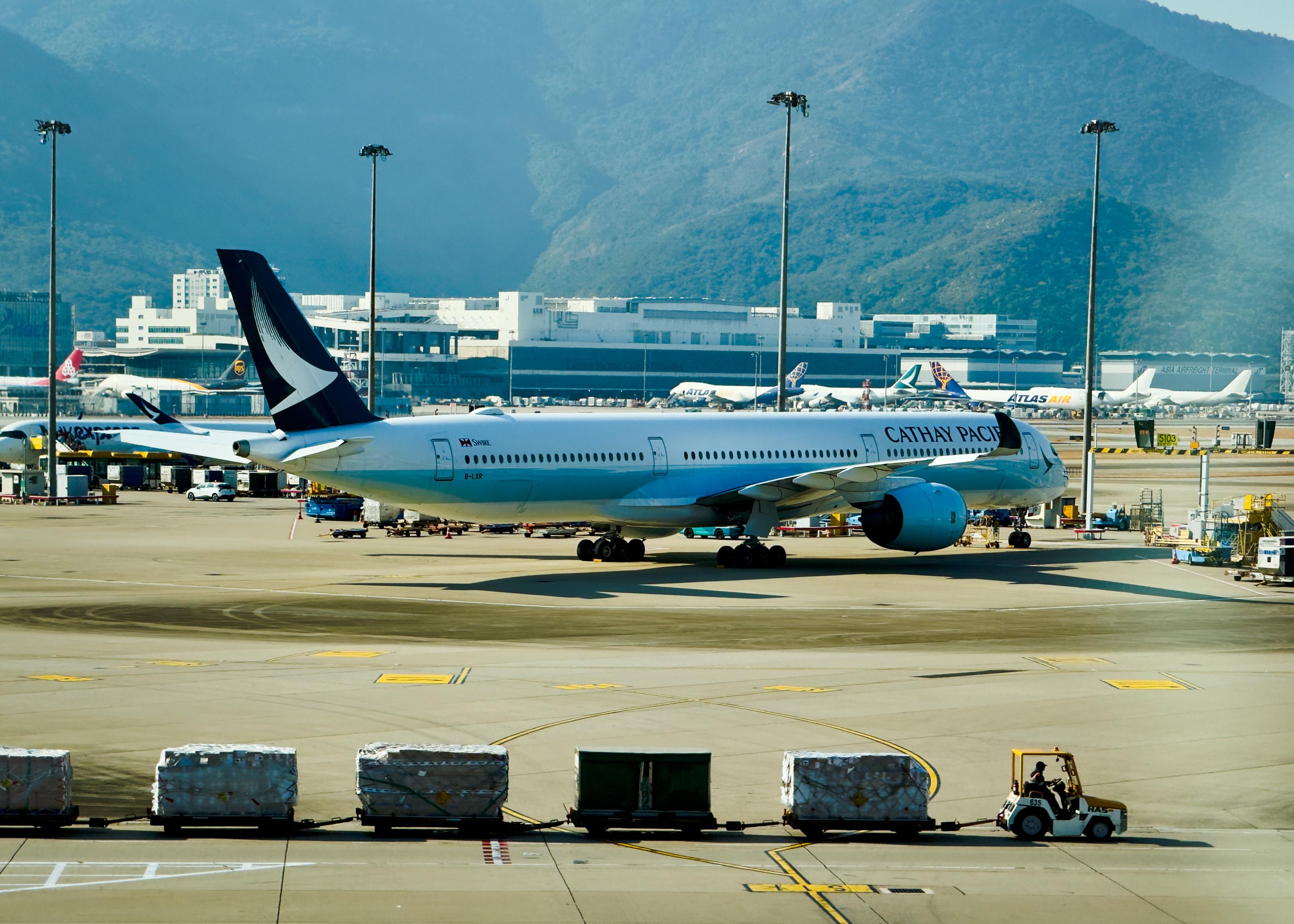 一架国泰航空的飞机在机场跑道上等待起飞，背景是山脉和其他飞机。