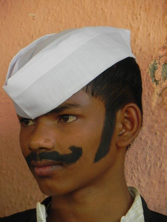 A young person in profile wears a white turban and face paint resembling a thick mustache and sideburns, standing against a textured orange wall.