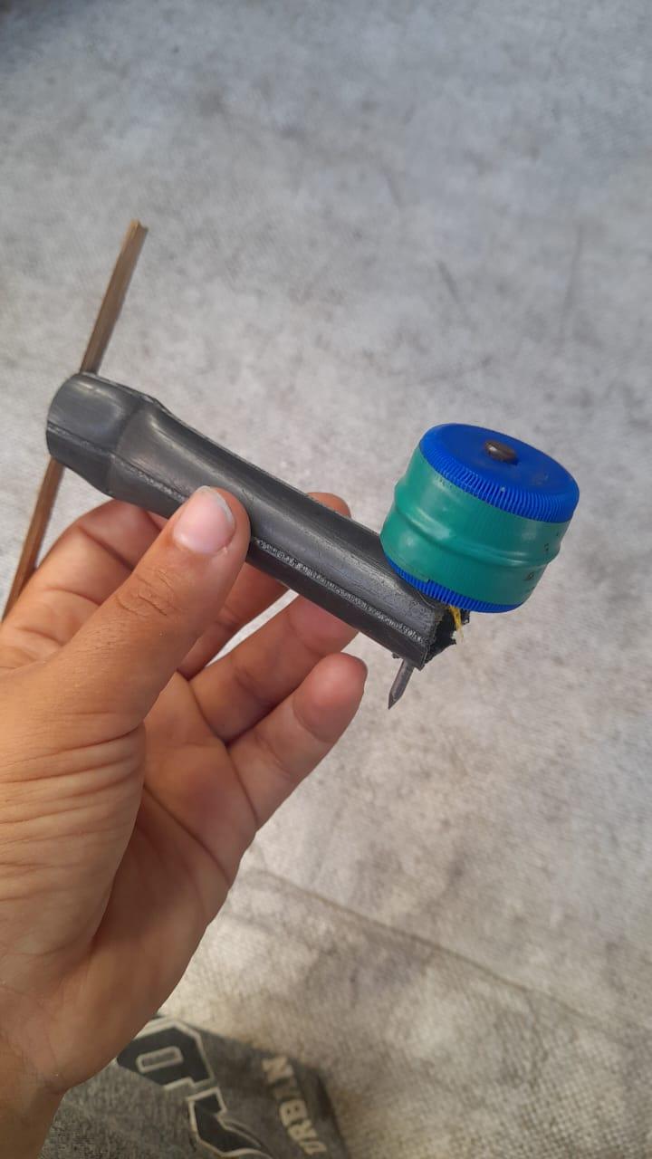 Hand holding a DIY spinning top made from a dark grey tube, a wooden stick, and a green and blue bottle cap with a nail.