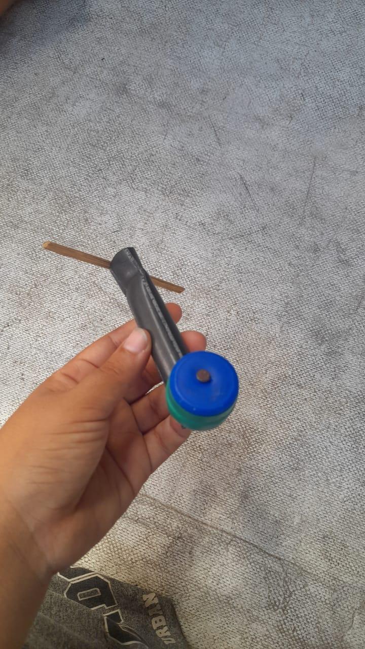 A handmade toy propeller, crafted from a piece of black tubing, a wooden stick, and two bottle caps, is held in a hand over a textured surface.