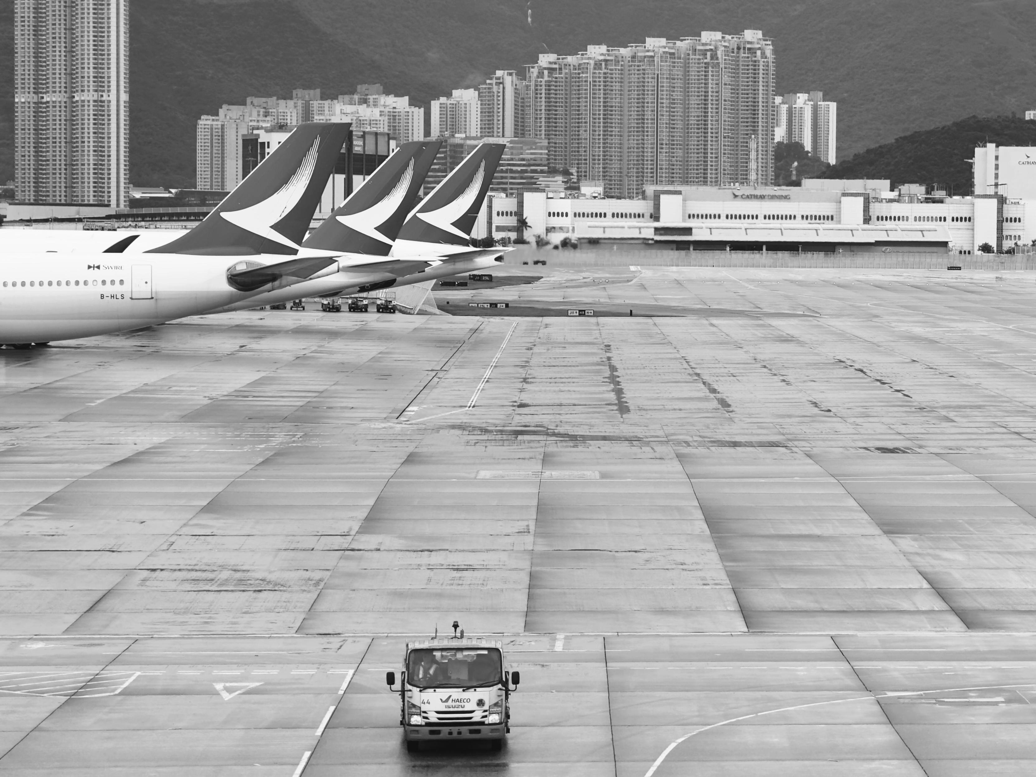 香港国际机场跑道上停着三架航空客机，背景是城市高楼和山峦。
