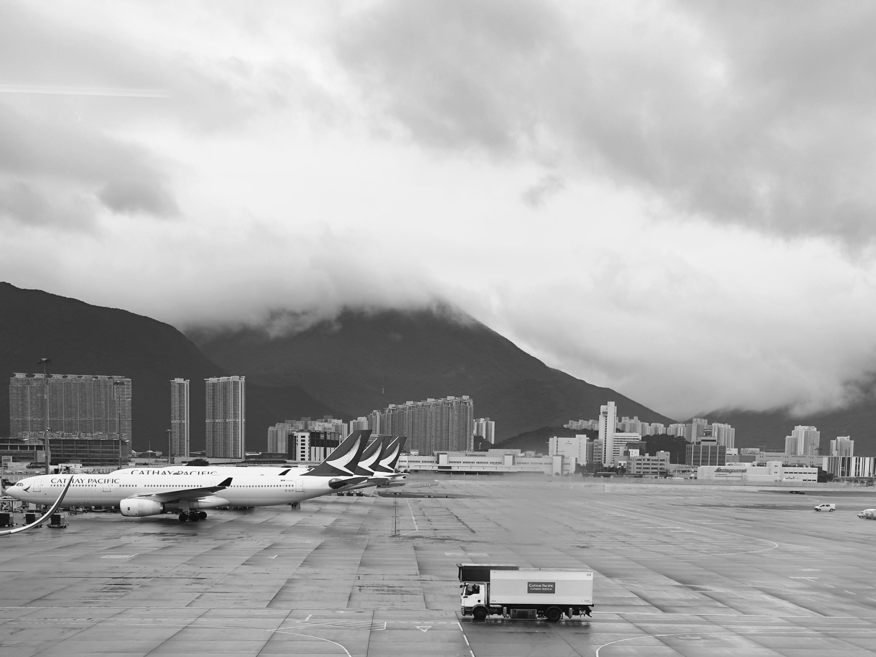 香港国际机场的跑道上停放着几架国泰航空的飞机，背景是高楼林立的城市和云雾缭绕的山峦。