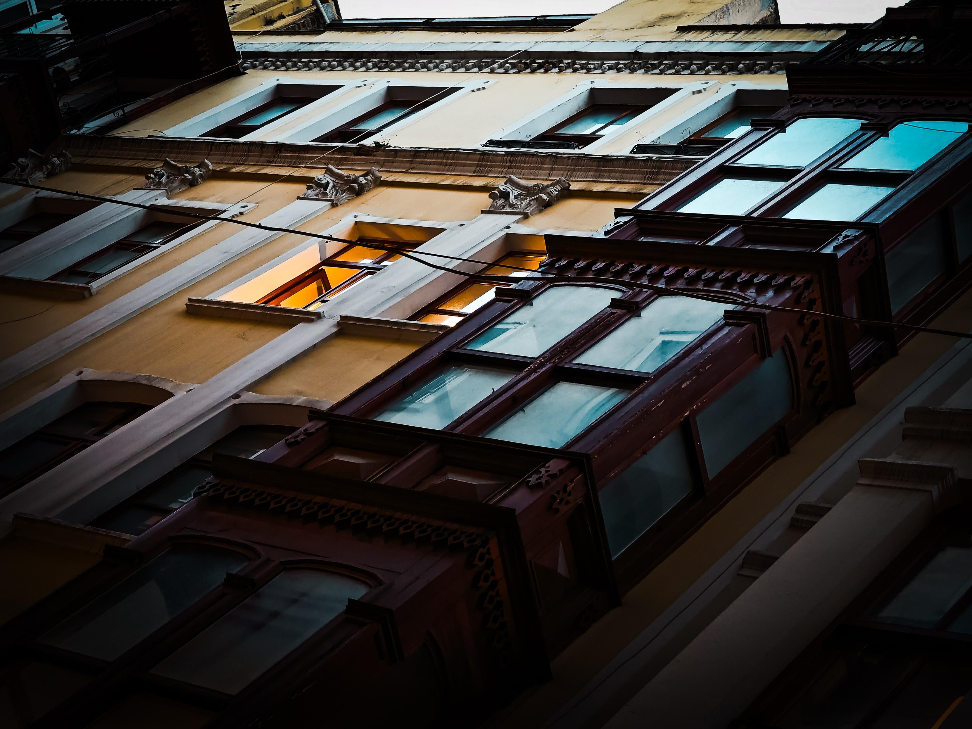 A photo taken at dusk looking up at the facia of a building with the windows reflecting the deepening blue sky as the sun goes down. Two open windows in a yellow building are illuminated from the inside. Beyoğlu, İstanbul, Türkiye