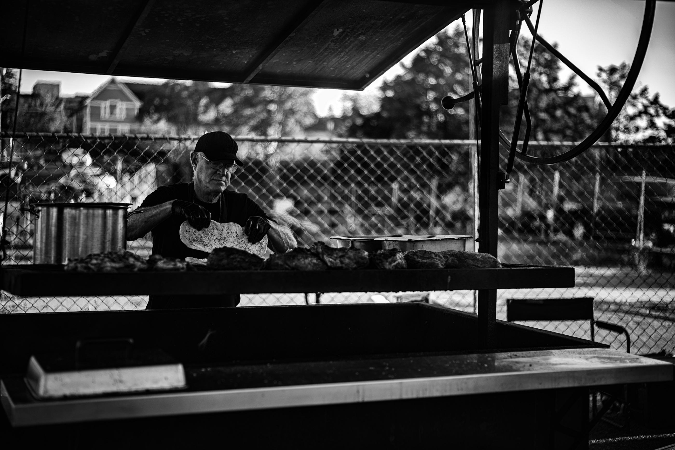 Black and white photo in portrait format of a man preparing orders over a mobile commercial charcoal grill.

The grill is covered by an adjustable wooden roof hydraulic roof with steel frame. The right centered supporting has a large diameter (~0.7m) circular grill height adjuster attached. A rectangular steel warming tray lid sits on the far left of the chrome colored table counter in front of the grill.

Eight large slabs of steak and chicken are lined from left to right cooking on the grill grate. A large stainless steel pot is in the far left corner simmering baked beans. Two medium sized rectangular warming trays are in the far right corner.

A man in his 60's wearing eyeglasses, and dressed in all black baseball cap, short sleeve T-shirt, apron, and latex gloves is pictured from waist up in the left center of the photo. He is holding a large hoagie in both hands above the grill ready to toast the opposite side.

The background consists of a chain link fence at the same height of 