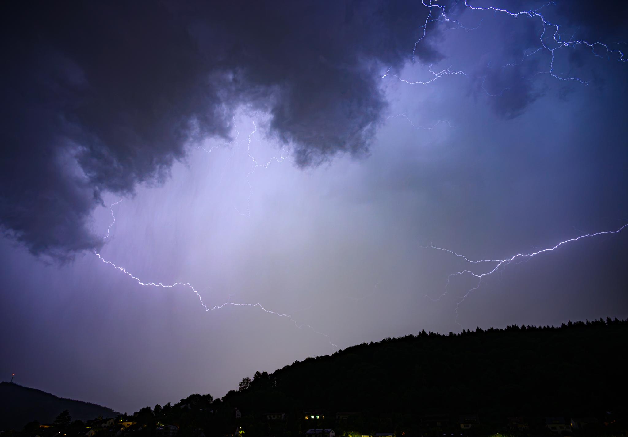 A night-time picture of the sky with a mostly dark hillside below, with a few lit houses. The sky is full of cloud, some darker above and to the left, illuminated by several bolts of lightning from the left and right sides. Most of the picture is blue grey, but the centre is a little more yellow, due to reflected streetlights from a city about 5km away.