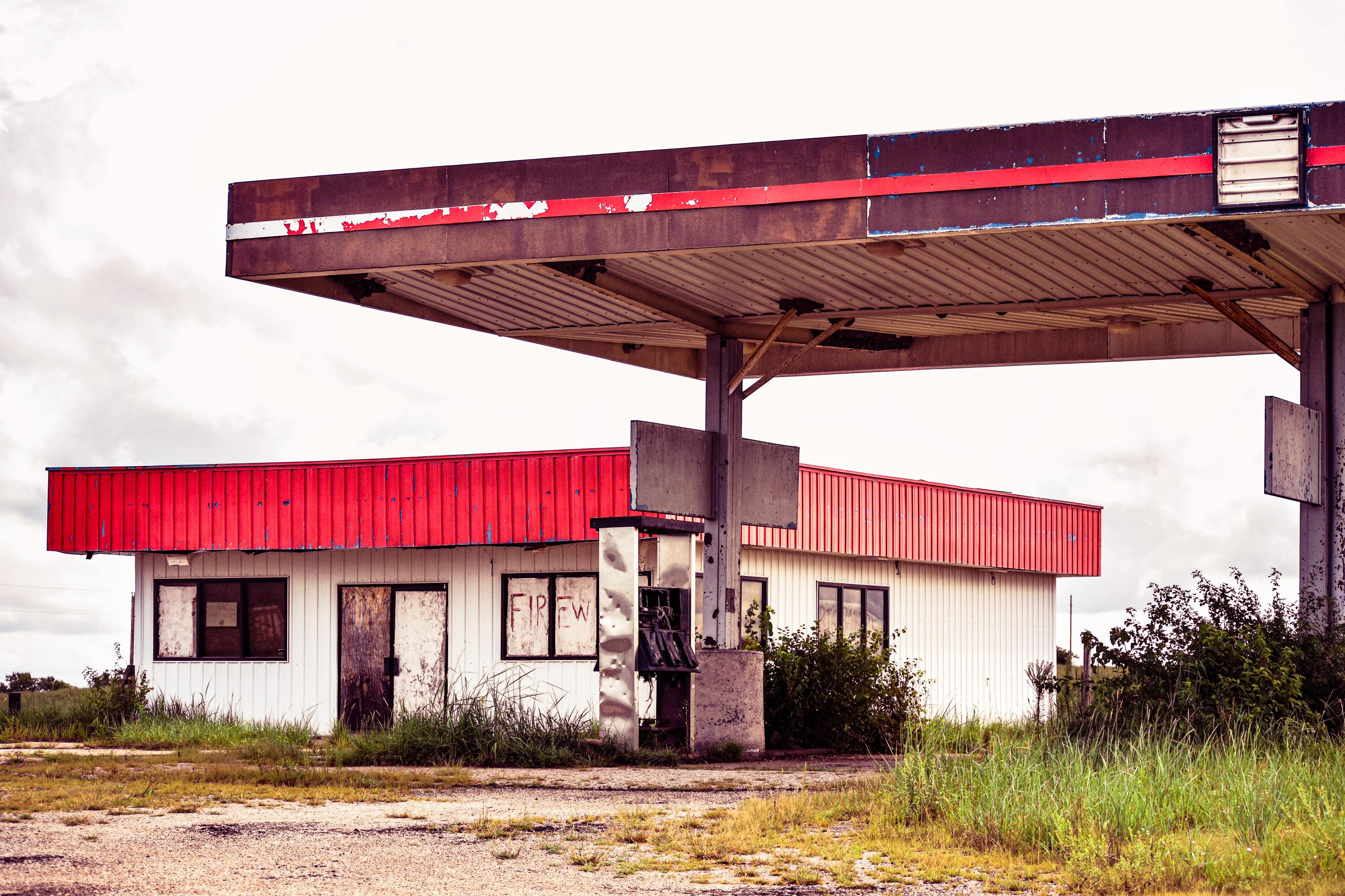 Abandoned gas station with a rusted red canopy, weathered white building, overgrown weeds, and cloudy skies creating a desolate mood.