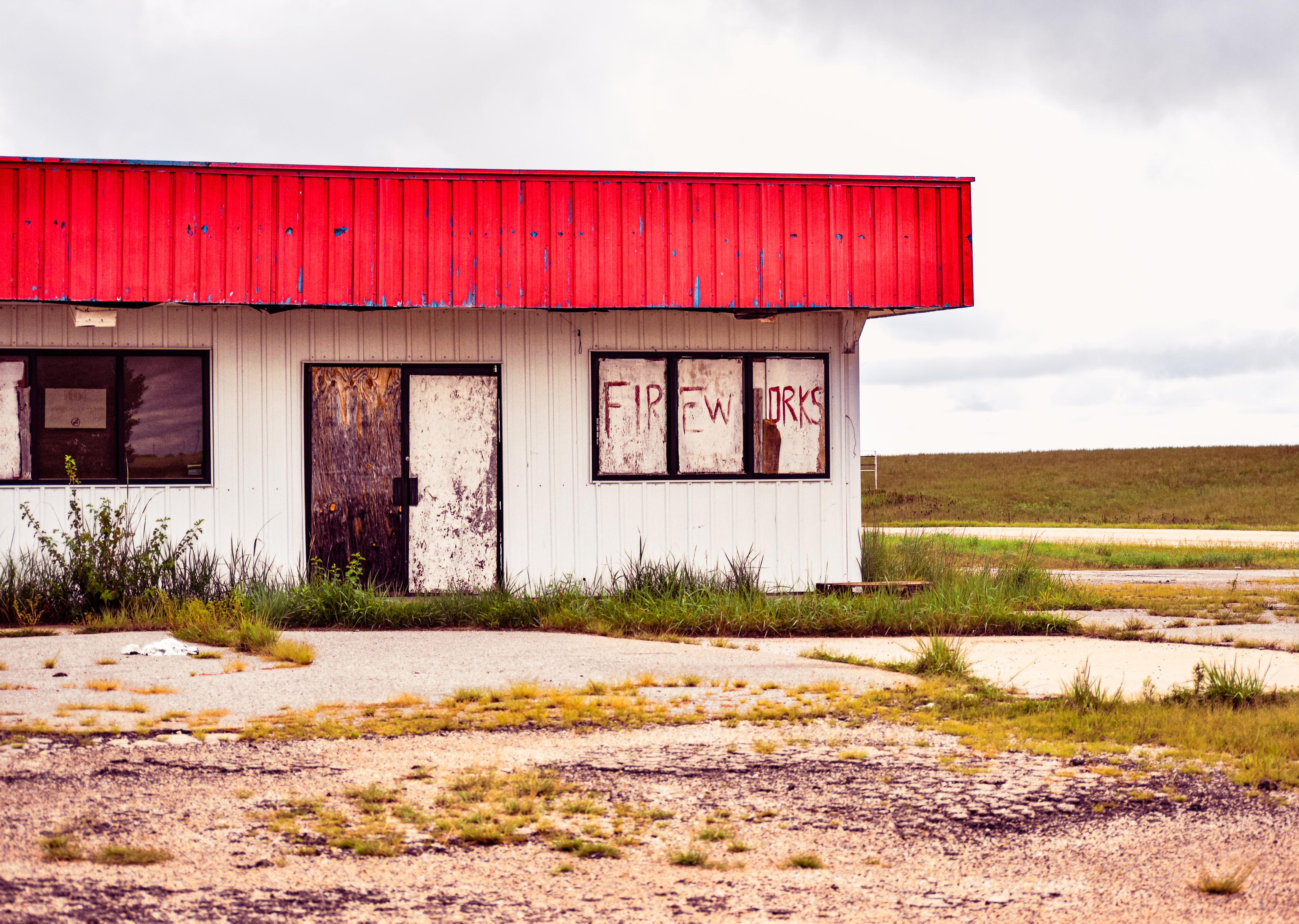 Abandoned rural building with a bright red roof, boarded windows, graffiti, and weeds amid cracked ground under an overcast sky.
