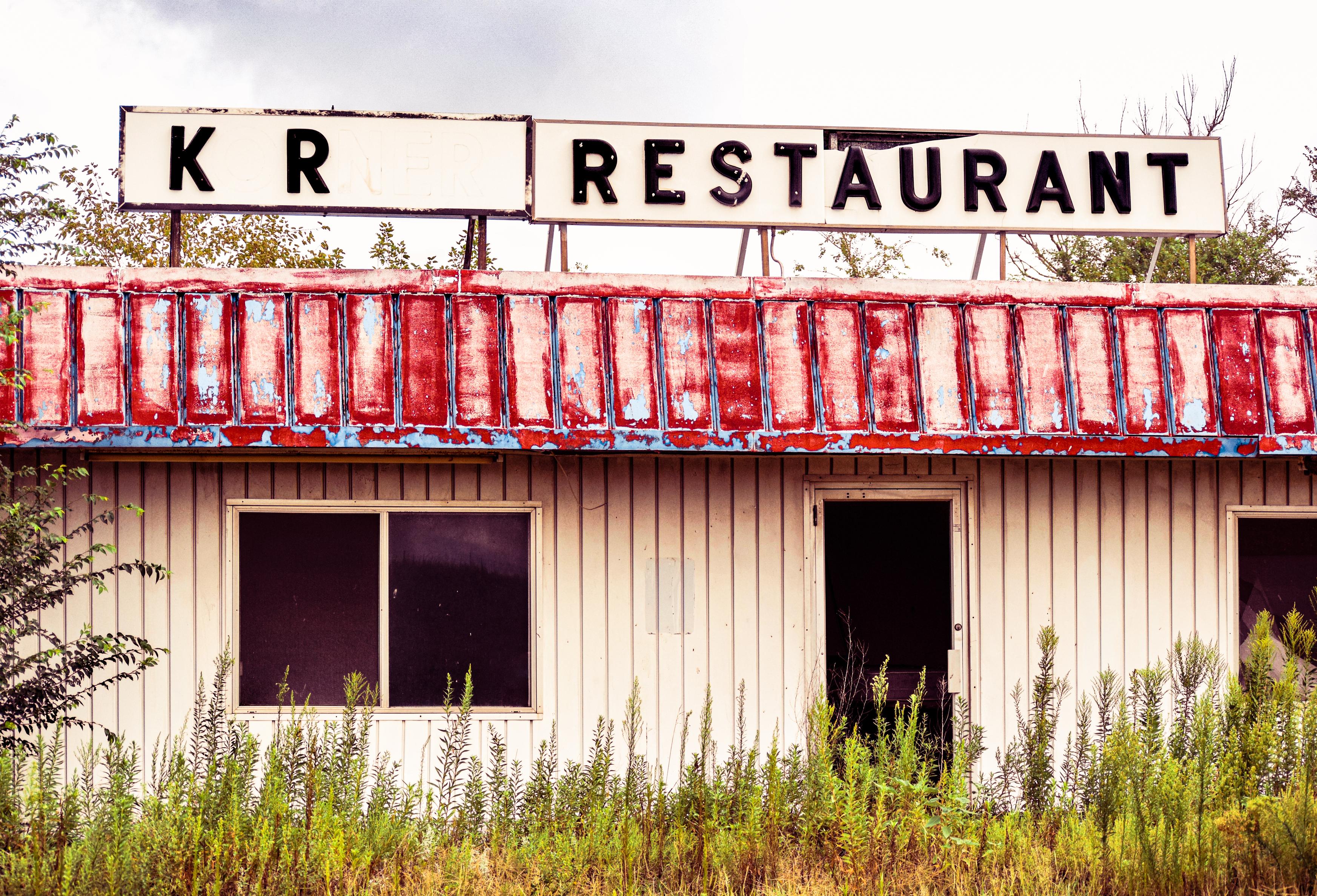 Weathered roadside “K R Restaurant” sign above a faded red awning, abandoned building, boarded windows, and overgrown weeds in daylight.