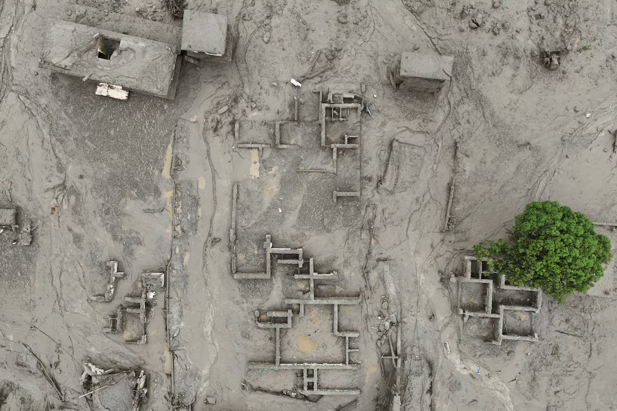 An aerial top-down view showing structures heavily submerged in thick grey mud following glacier collapse. Only the upper outlines of roofless walls, foundation footprints, and concrete buildings remain visible amidst the vast layer of sludge, with a single vibrant green tree standing on the right side.