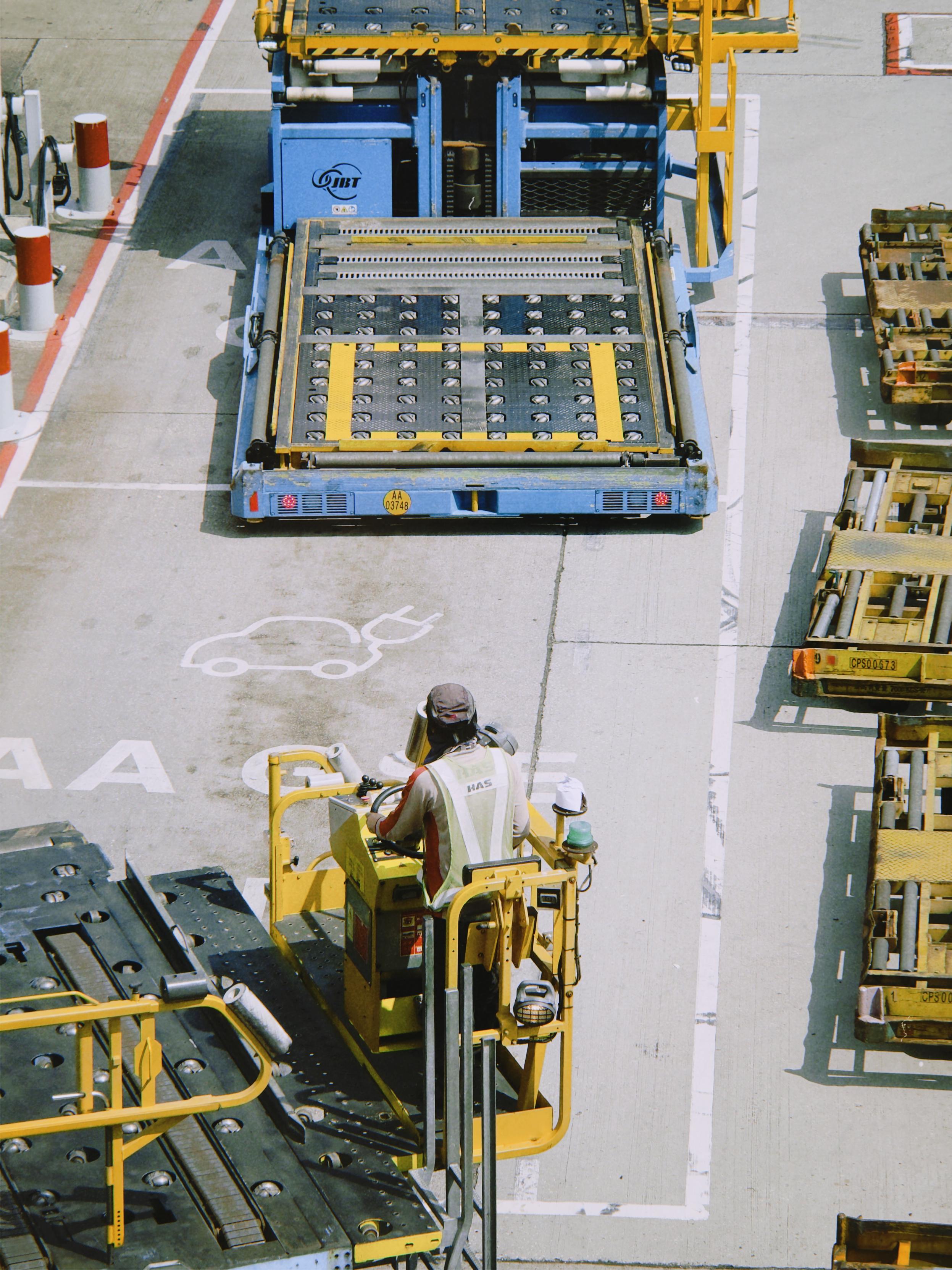 Worker operating a yellow electric vehicle near a blue vehicle charger in an outdoor parking lot.