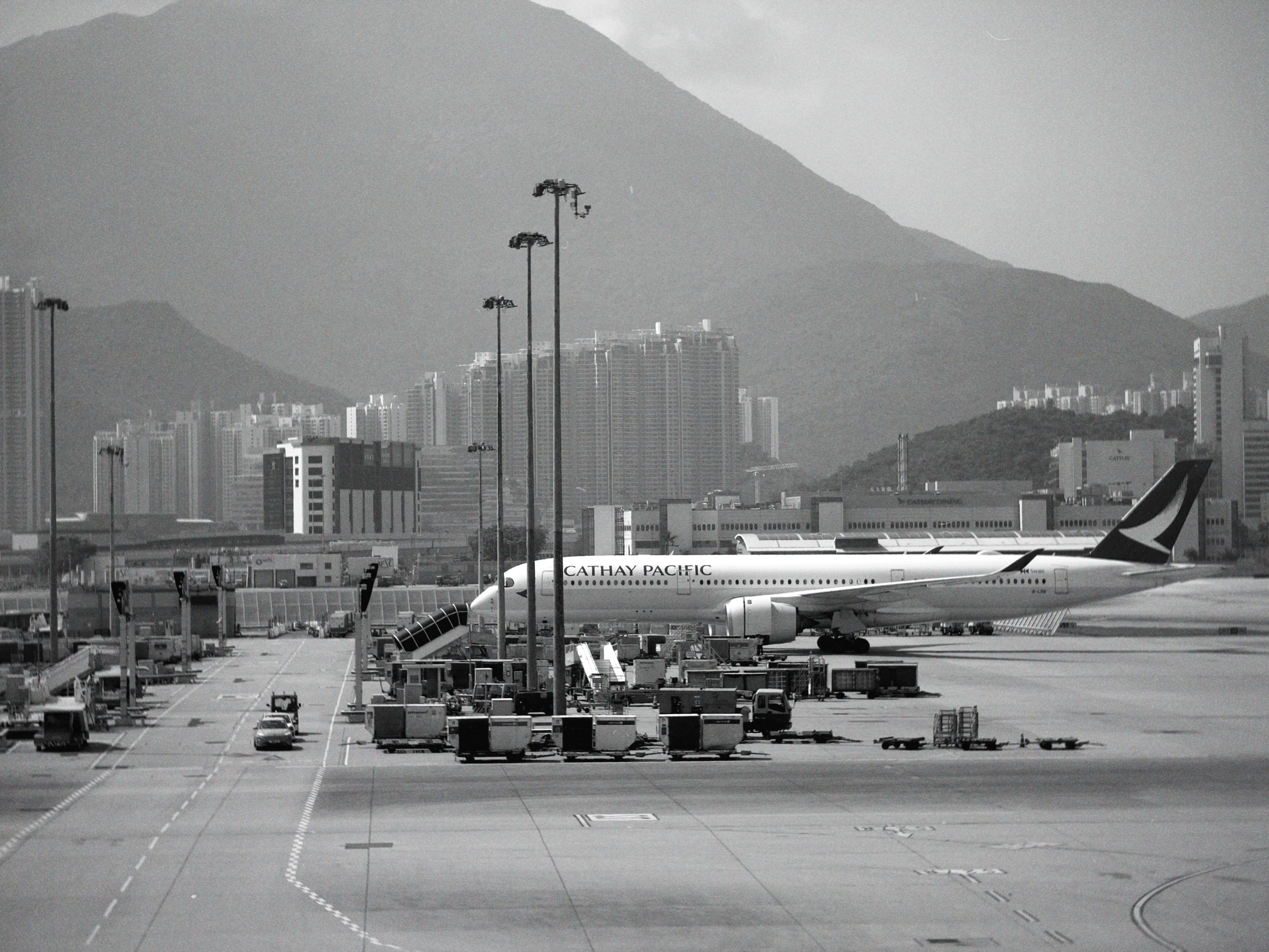 飞机停靠在香港国际机场，背景是高楼和山脉，画面为黑白色调。