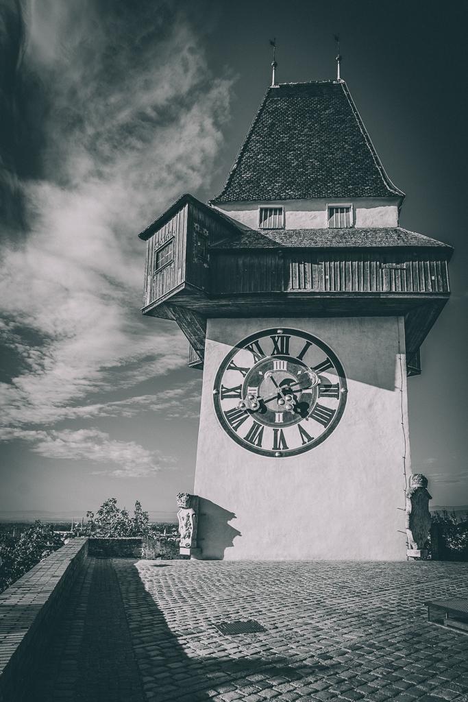 Eine Schwarz-Weiß-Fotografie im Hochformat zeigt den Grazer Uhrturm auf dem Schlossberg. Der Turm hat ein großes Zifferblatt mit römischen Ziffern und eine markante Holzgalerie, die über die Turmmauer hinausragt. Er steht auf einer gepflasterten Aussichtsterrasse. Der Himmel ist dramatisch mit Wolken durchsetzt, und im Hintergrund ist ein Blick auf die Stadt Graz zu erkennen.