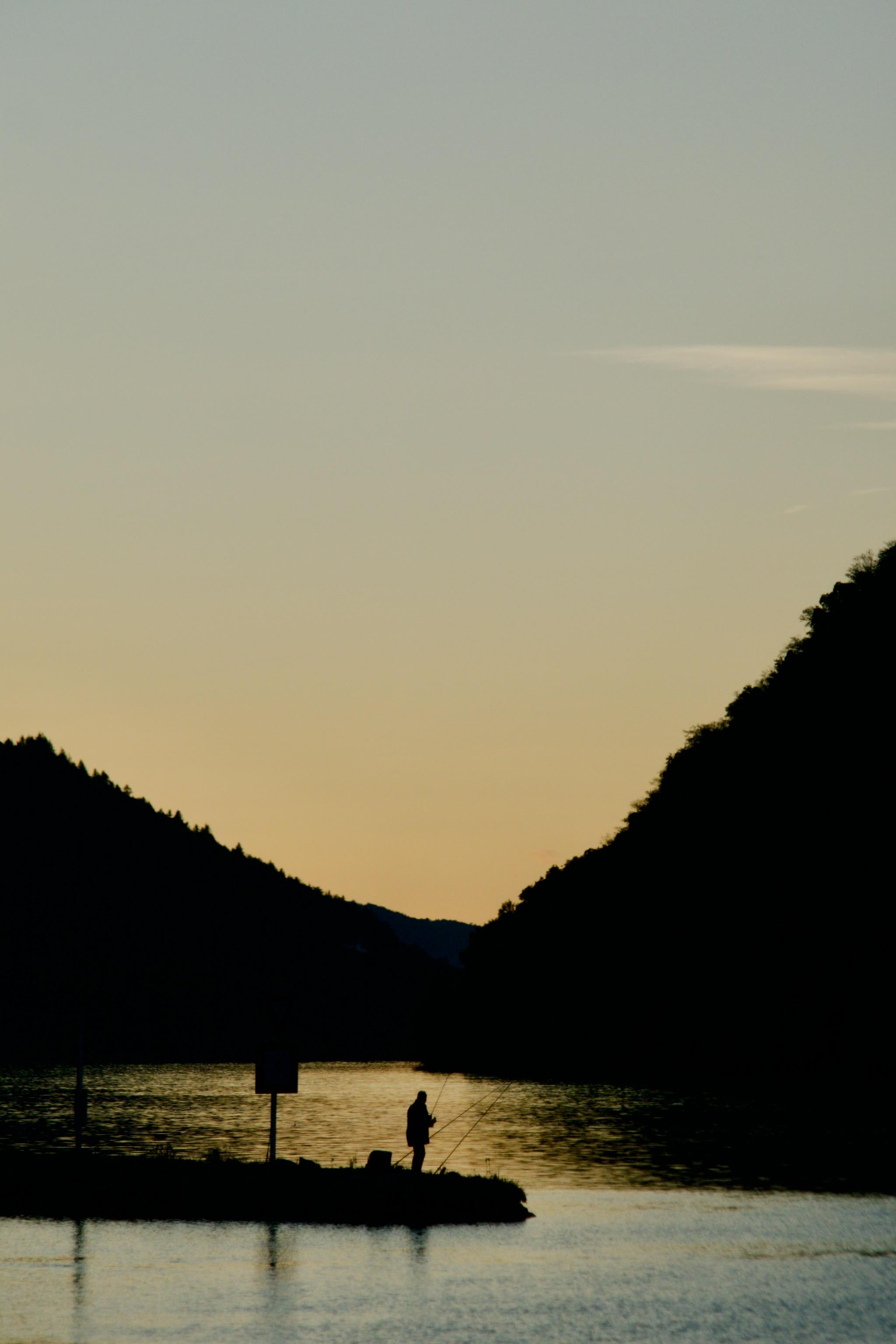 Picture of the river Danube after the sun went down.The surrounding hills are black triangles with uneven edges because of the trees. In the middle lower part of the picture is a little island and a man standing on the waterfront and fishing. 
The river band is flowing in curves, the colour is changing from dark orange, brownish in the background to blue in the foreground.