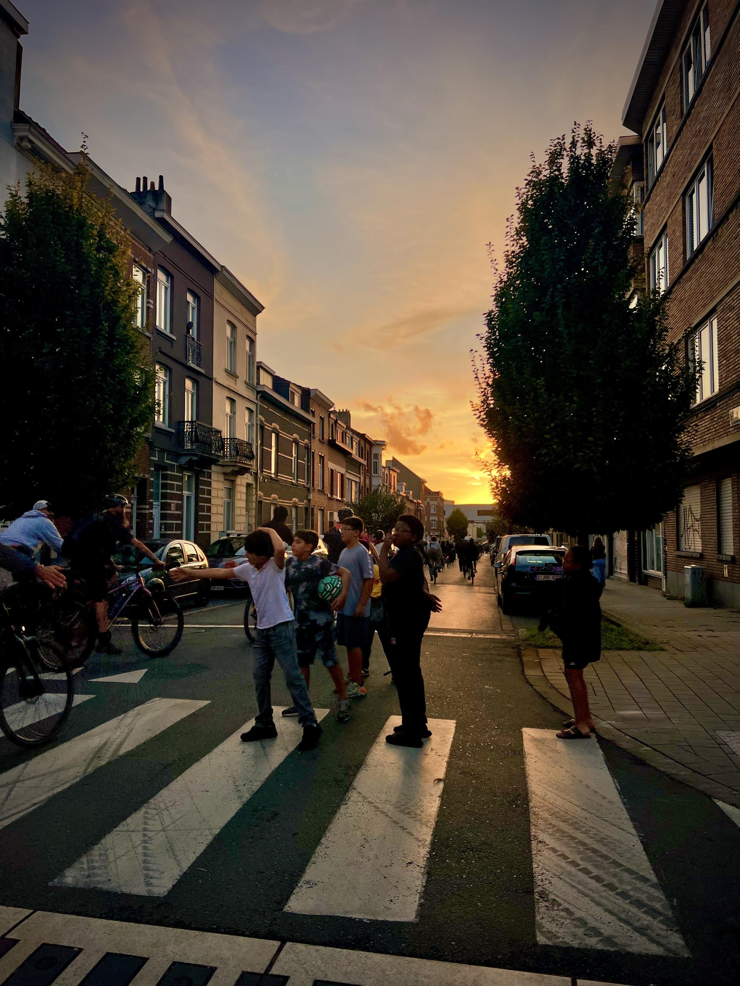 Notte Luc J.g, Vorst/Forest, Brussels-Capital, Belgium

Children play on a zebra crossing at sunset in an urban neighborhood with brick buildings lining both sides of the street. The road is paved asphalt, and trees line the sidewalks. A few bicycles are parked nearby, and people walk along the street. The sky is a gradient from orange to blue as the sun sets behind distant buildings.

35 mm equivalent: 26 mm