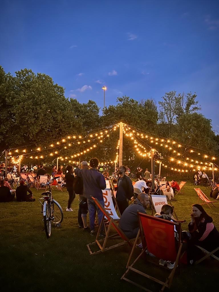 La Guinguette des Etangs, Anderlecht, Brussels-Capital, Belgium

A lively outdoor gathering under a canopy of string lights strung between wooden poles on a grassy field at dusk. People are seated in red deck chairs and standing among bicycles, enjoying the evening. The sky is clear with a few scattered clouds above dense trees. 'APEROL' text appears on some signs.

35 mm equivalent: 26 mm