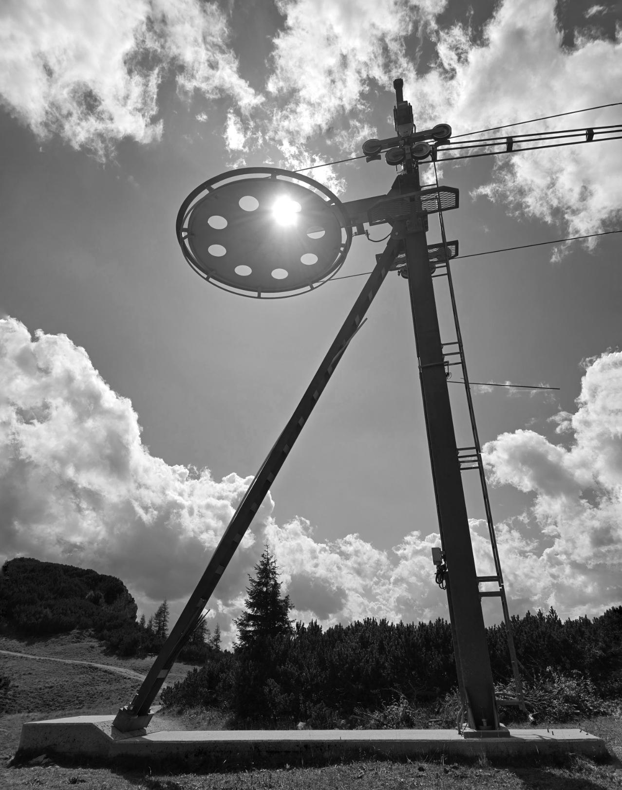 Schwarzweiß. Auf dem Berg. Sonne. Teilweise bewölkt. Ein Luftseilbahn-Mast. Durch ein Loch des Mastes scheint die Sonne.