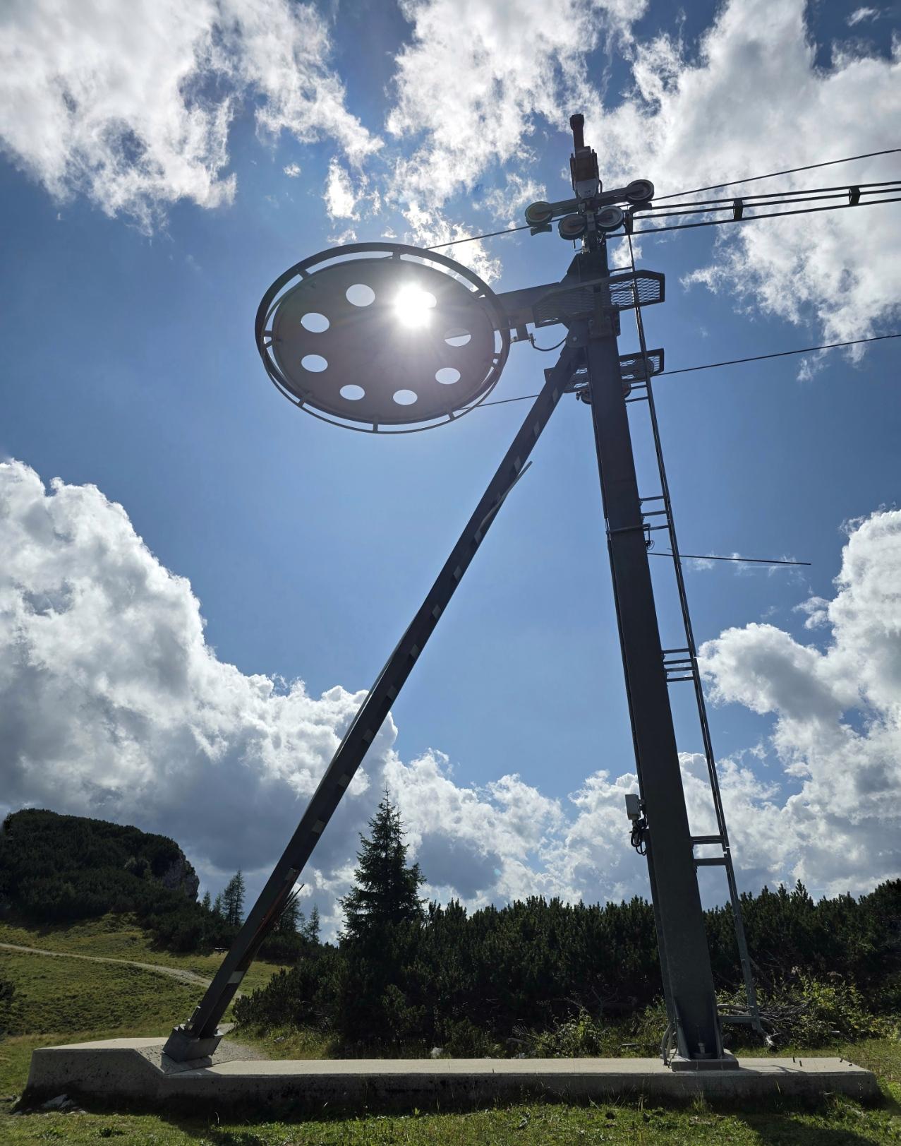 Farbig. Auf dem Berg. Sonne. Teilweise bewölkt. Ein Luftseilbahn-Mast. Durch ein Loch des Mastes scheint die Sonne.