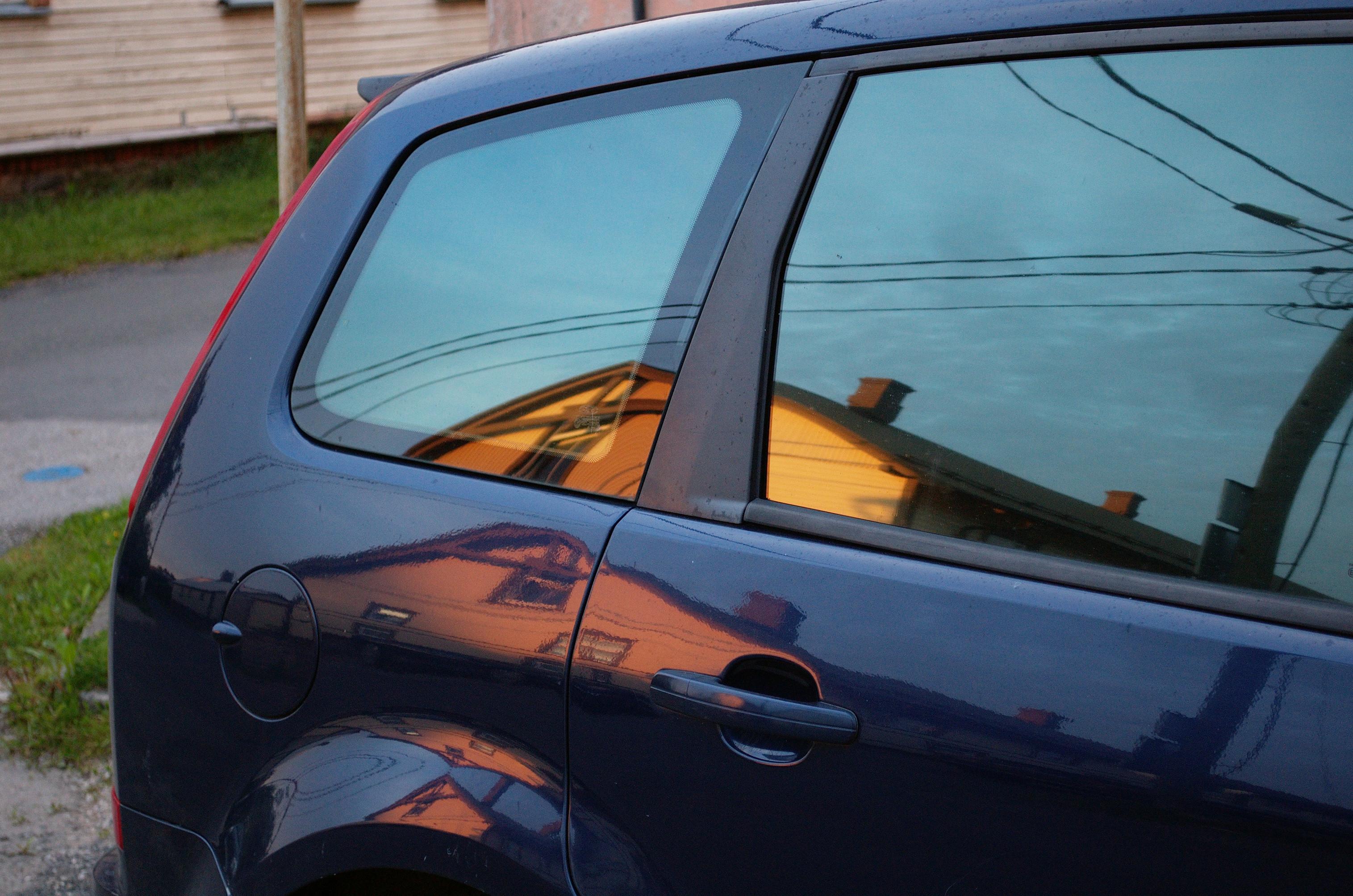 Close-up photo of the side of a dark blue car, showing reflections of a yellow wooden house on the other side of the street. The reflection on the windows is yellow, another partial reflection on the body looks more red, and the curved edge of the wheel well shows two very distorted reddish reflections, one of them upside down.