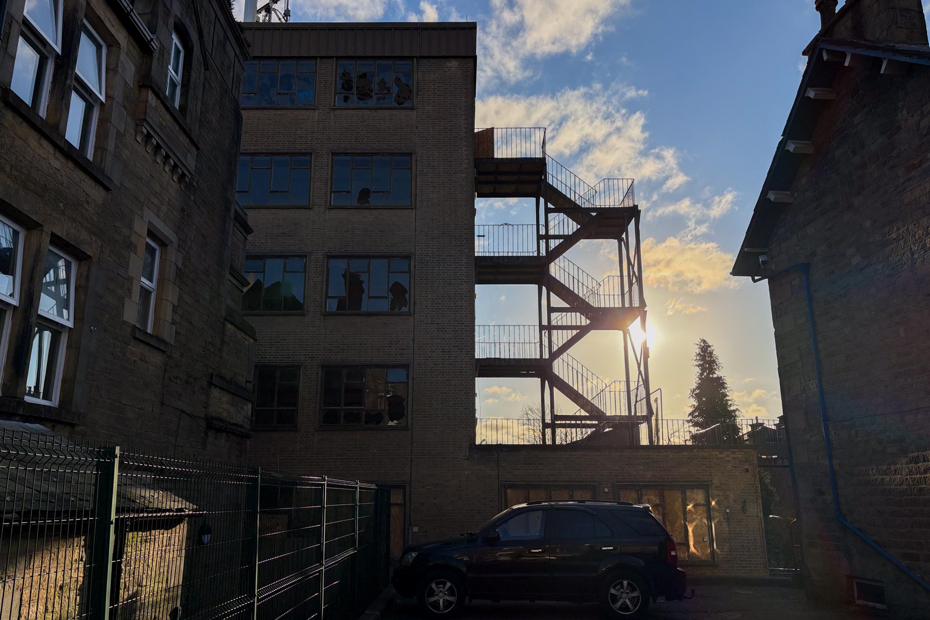 A black SUV is parked beside a fenced building. The sun is setting behind a brick building with metal staircases. Shadows and reflections are visible on the windows, and the sky is partly cloudy with yellow and blue hues.