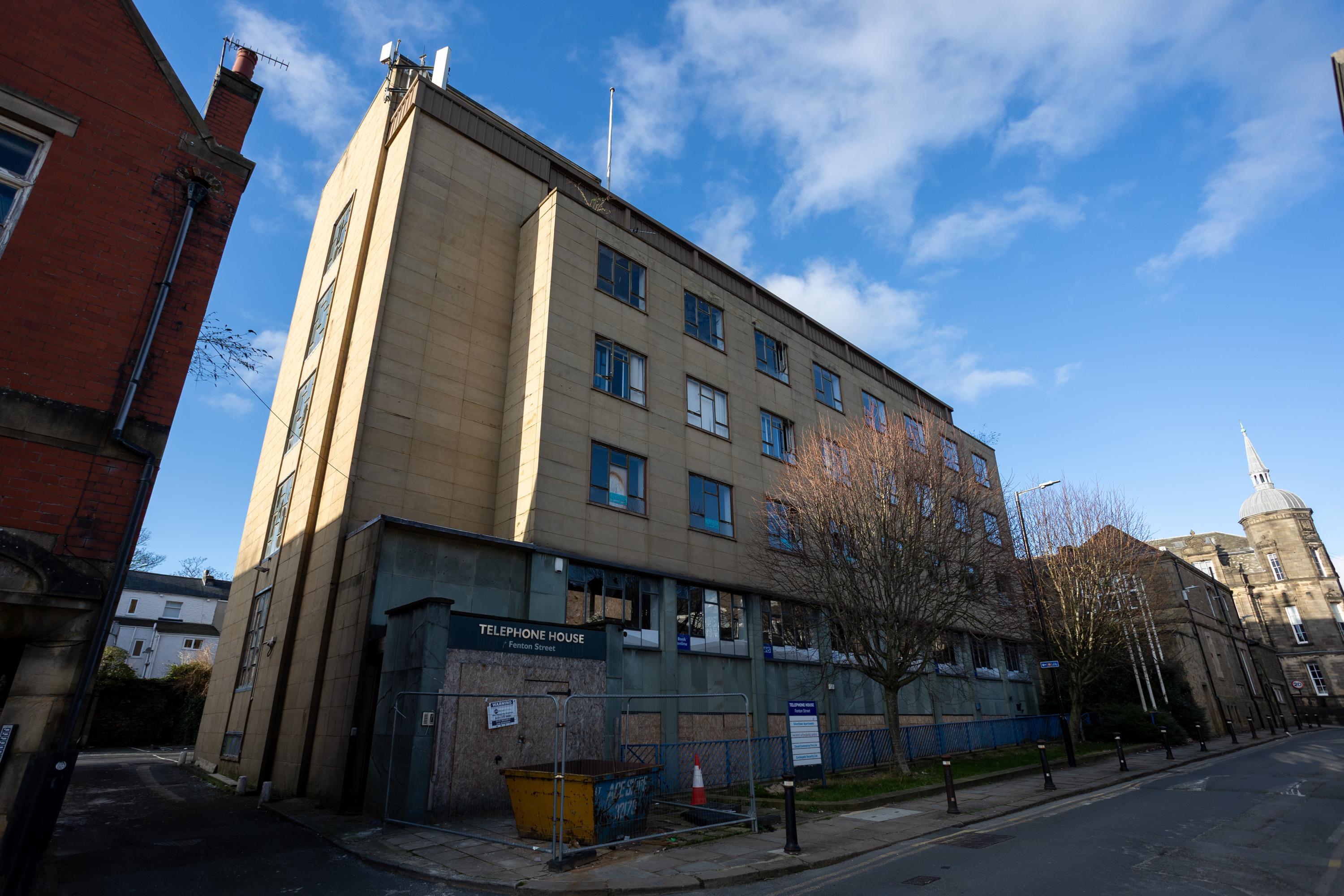 A boarded-up, beige four-storey office building labelled TELEPOINT HOUSE stands on a quiet street, with a yellow skip in front. The windows are broken or missing, and leafless trees line the pavement. The sky is partly cloudy and blue.