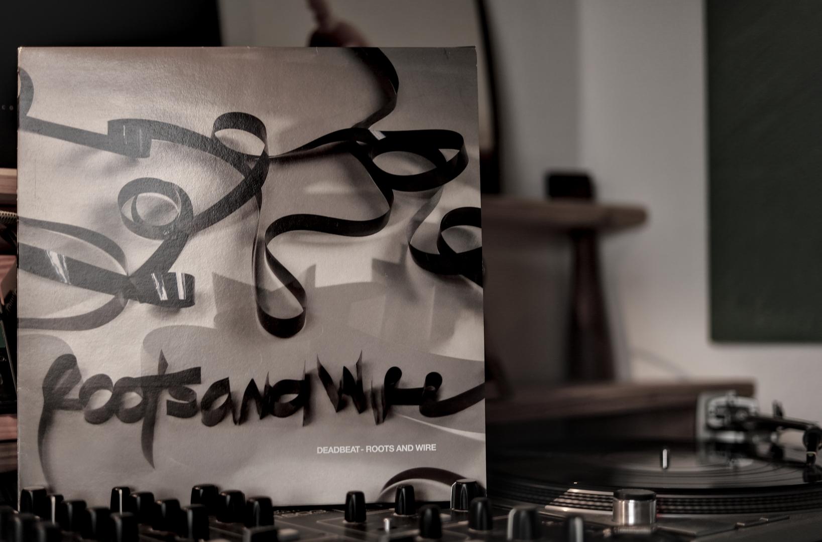 Deadbeats' “Roots and Wire” album, the cover, is sitting on a mixer. To the right of it is a turntable. The background is out of focus. The focus is clearly on the cover.
