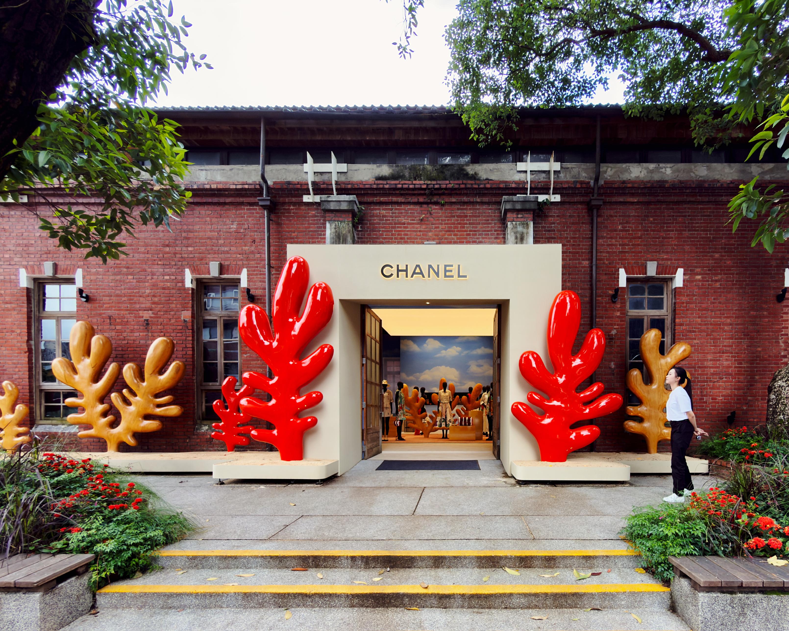Chanel门店外观，装饰有红色和金色树形雕塑，建筑为红砖墙，有两扇开启的门，店内背景为蓝天白云。