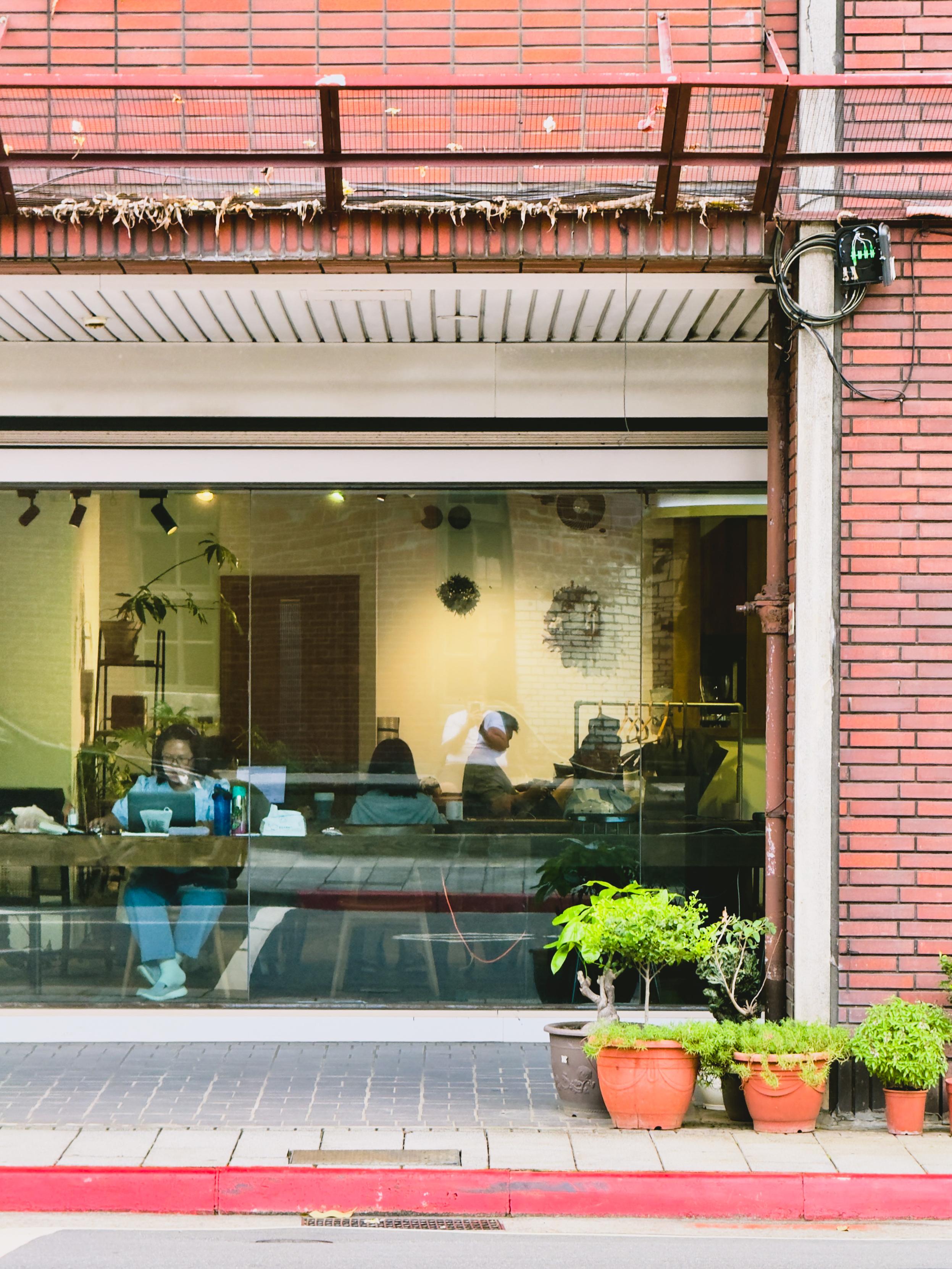 People working inside a cafe, seen through a large glass window, with plants outside on a sidewalk.
