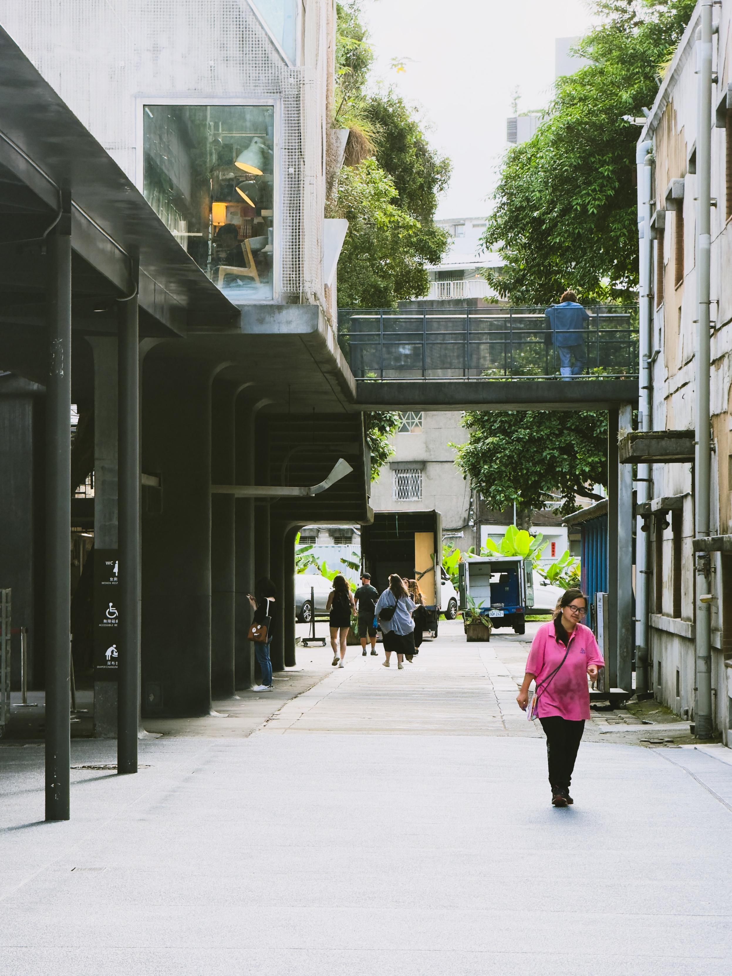 People walking on a city sidewalk with modern buildings and greenery, during daytime.