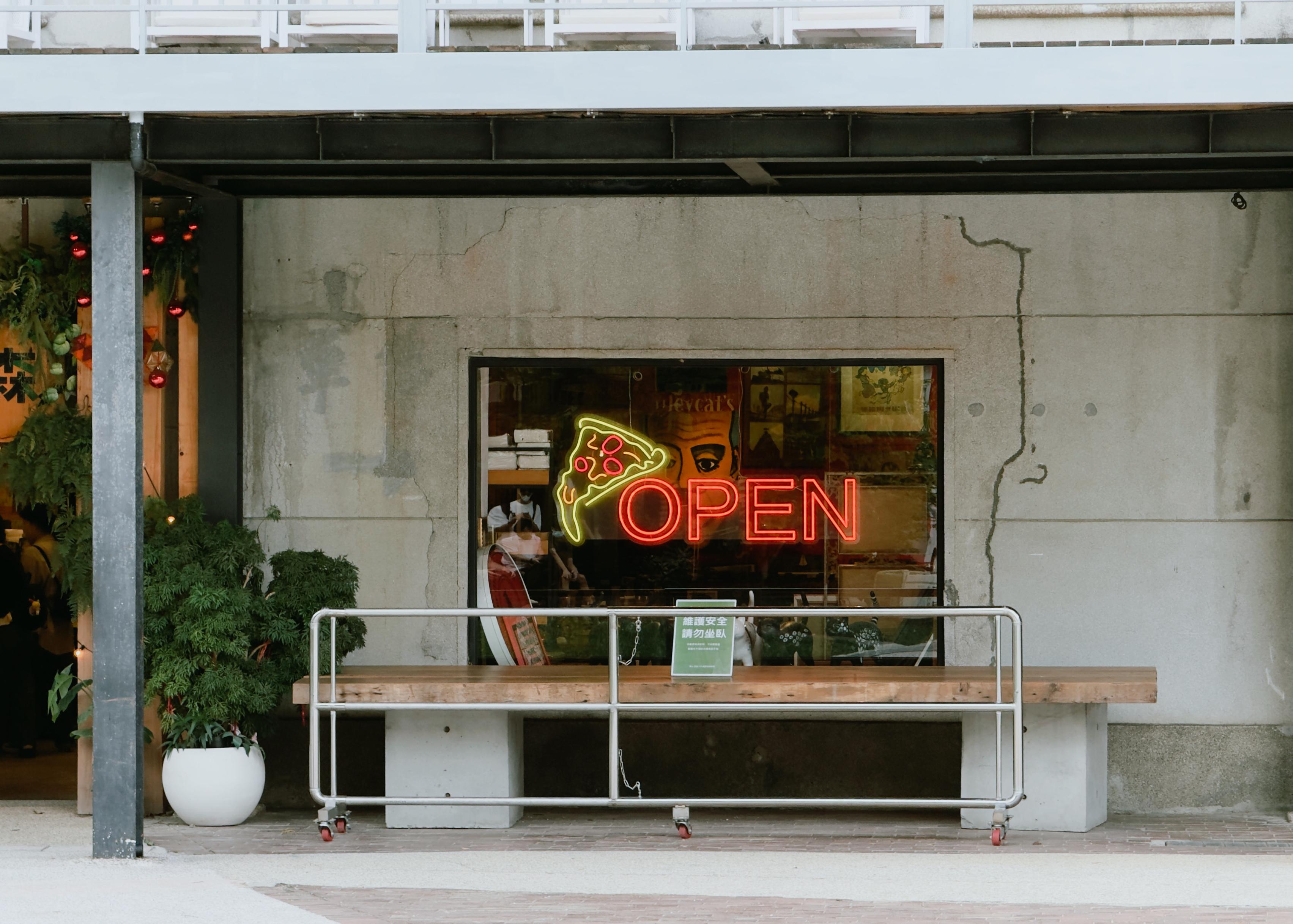 A storefront window with a neon "OPEN" sign and a pizza slice graphic, surrounded by a concrete wall and a small bench outside.