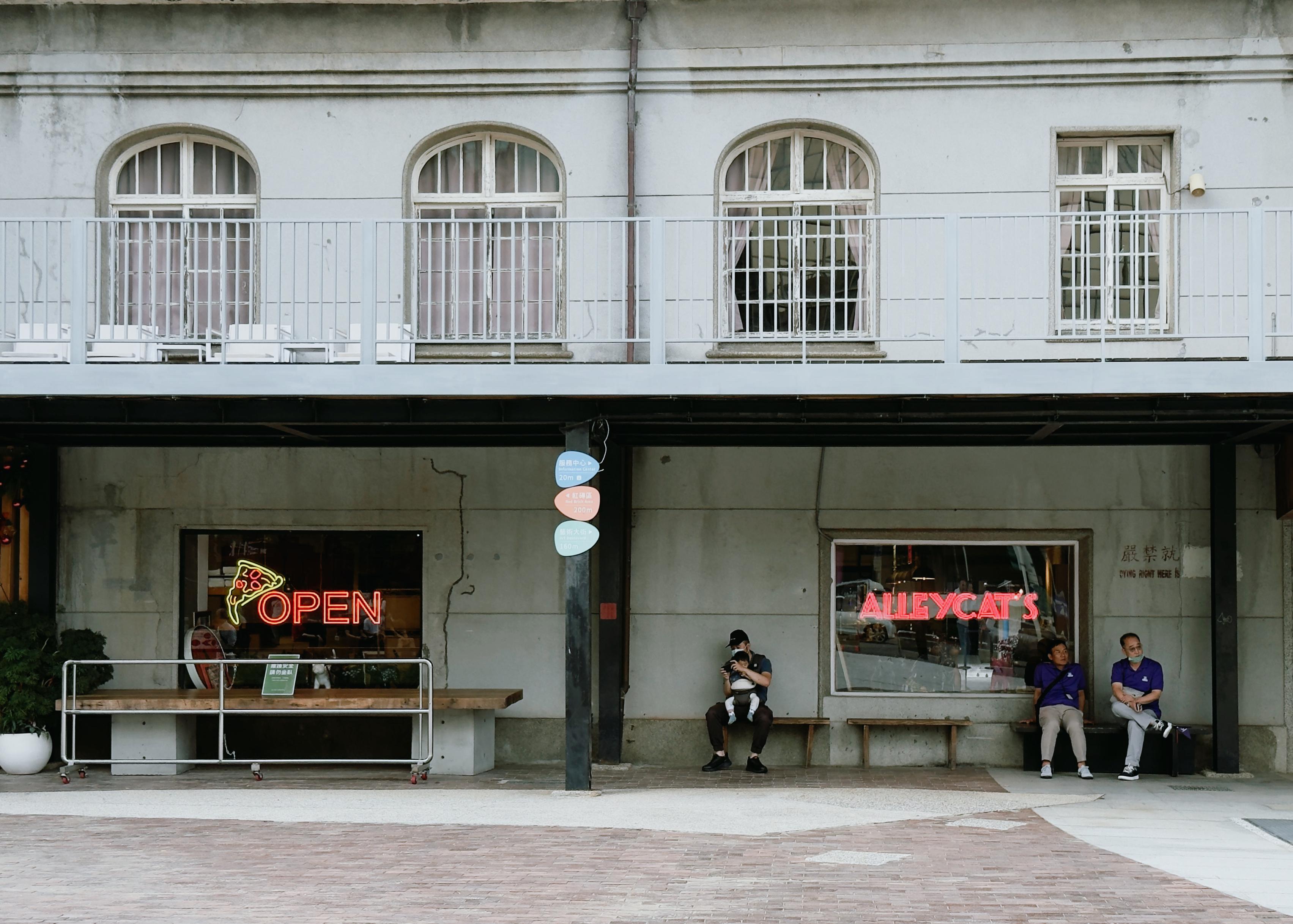 People sitting outside a storefront with neon signs reading "OPEN" and "ALLEYCAT'S". One person is standing, two are sitting on a bench, and one is using a cellphone.