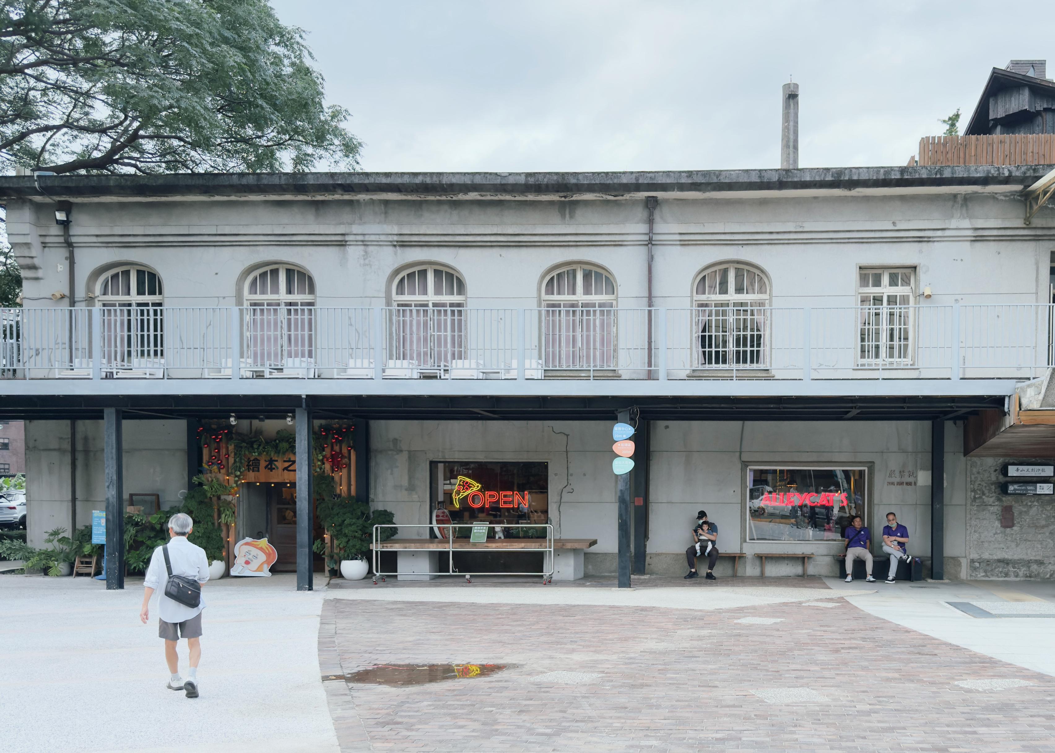 People sitting outside a cafe on a street with a person walking away in the foreground. The cafe has "OPEN" and "ALLEY CATS" signs in the window.