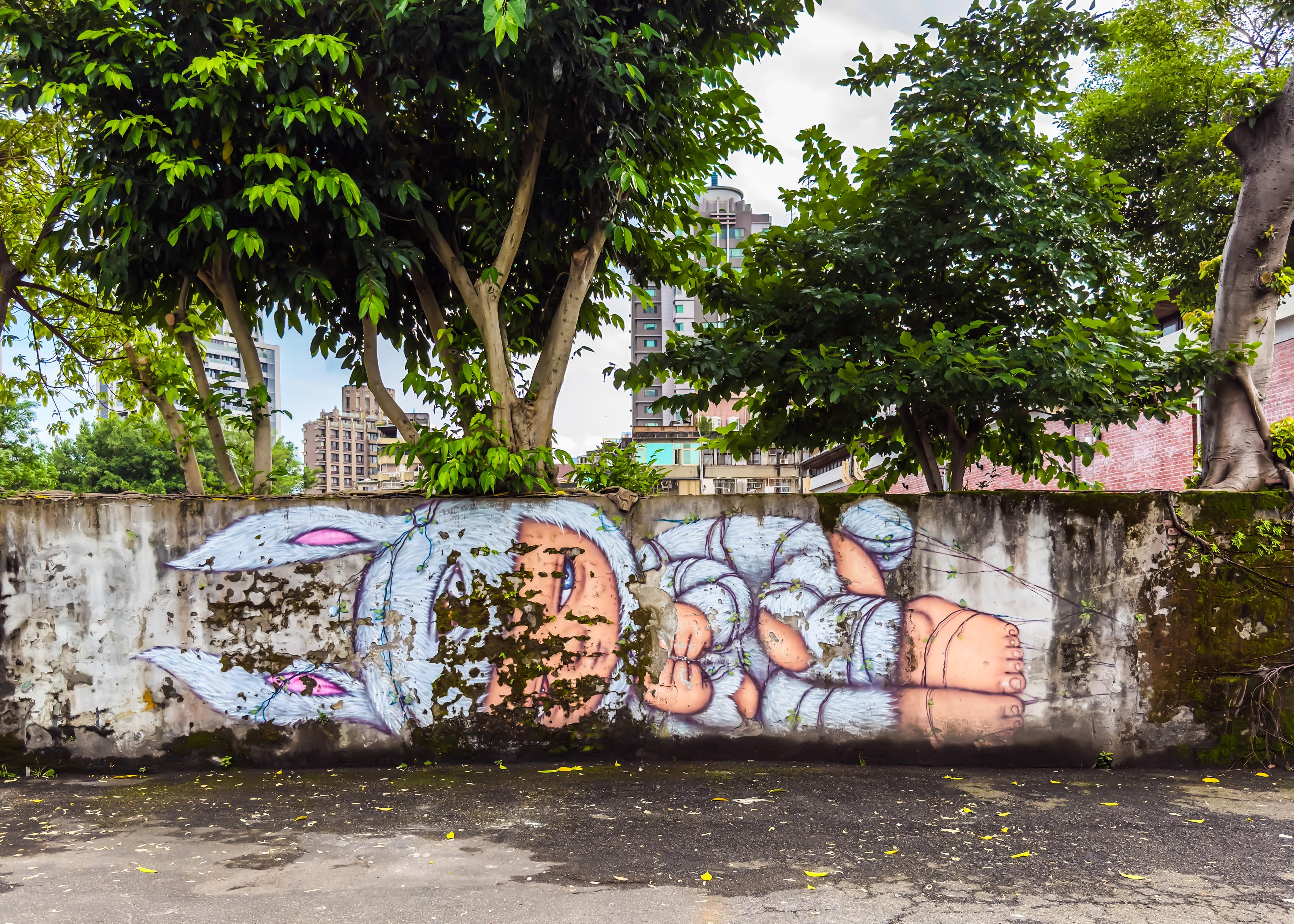 Colorful mural of a sleeping child with a bunny hat on a weathered concrete wall, surrounded by trees and city buildings in the background.