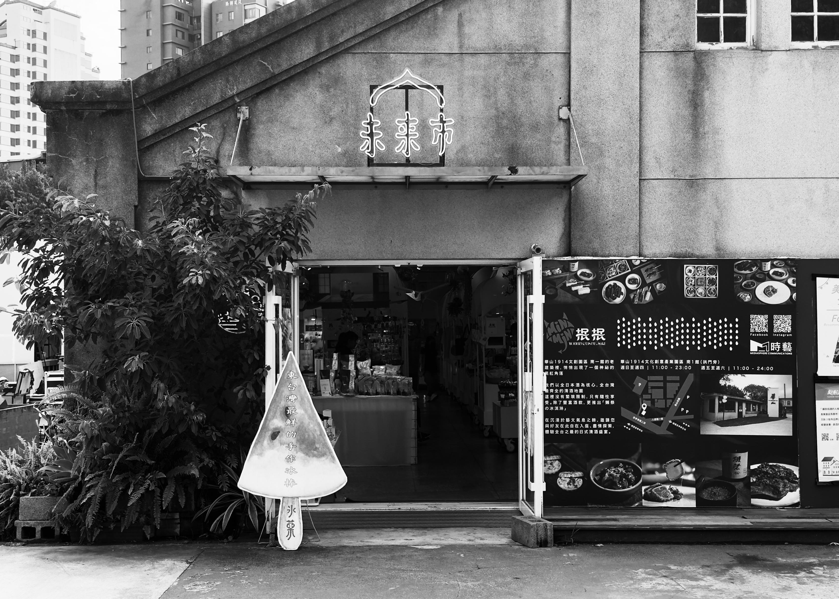 Black and white photo of a small shop with open doors, a sign with Chinese characters above the entrance, and a large menu board outside.