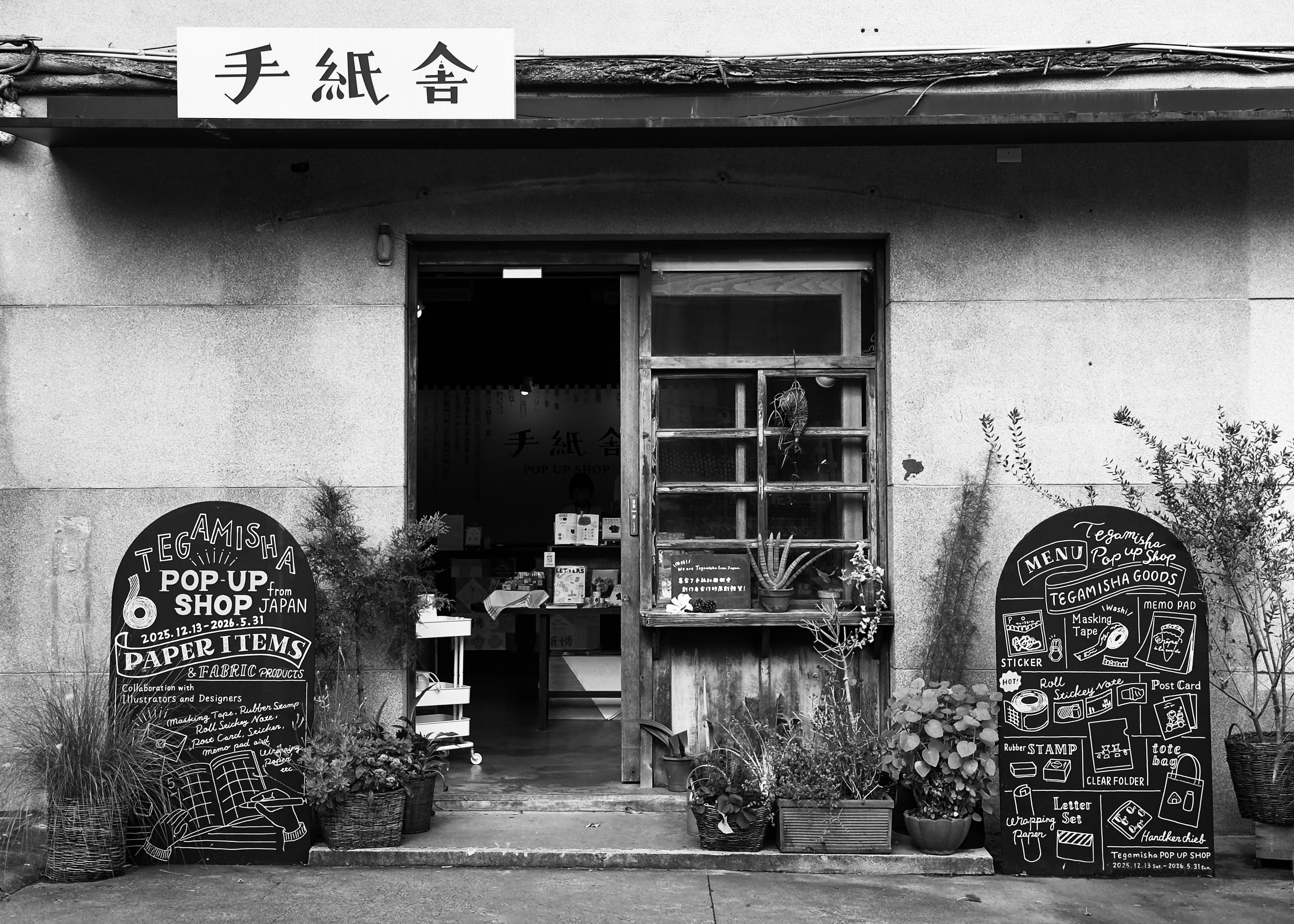 Black and white storefront with handwritten signs, potted plants, and a partially open door, promoting a pop-up shop in Japan.