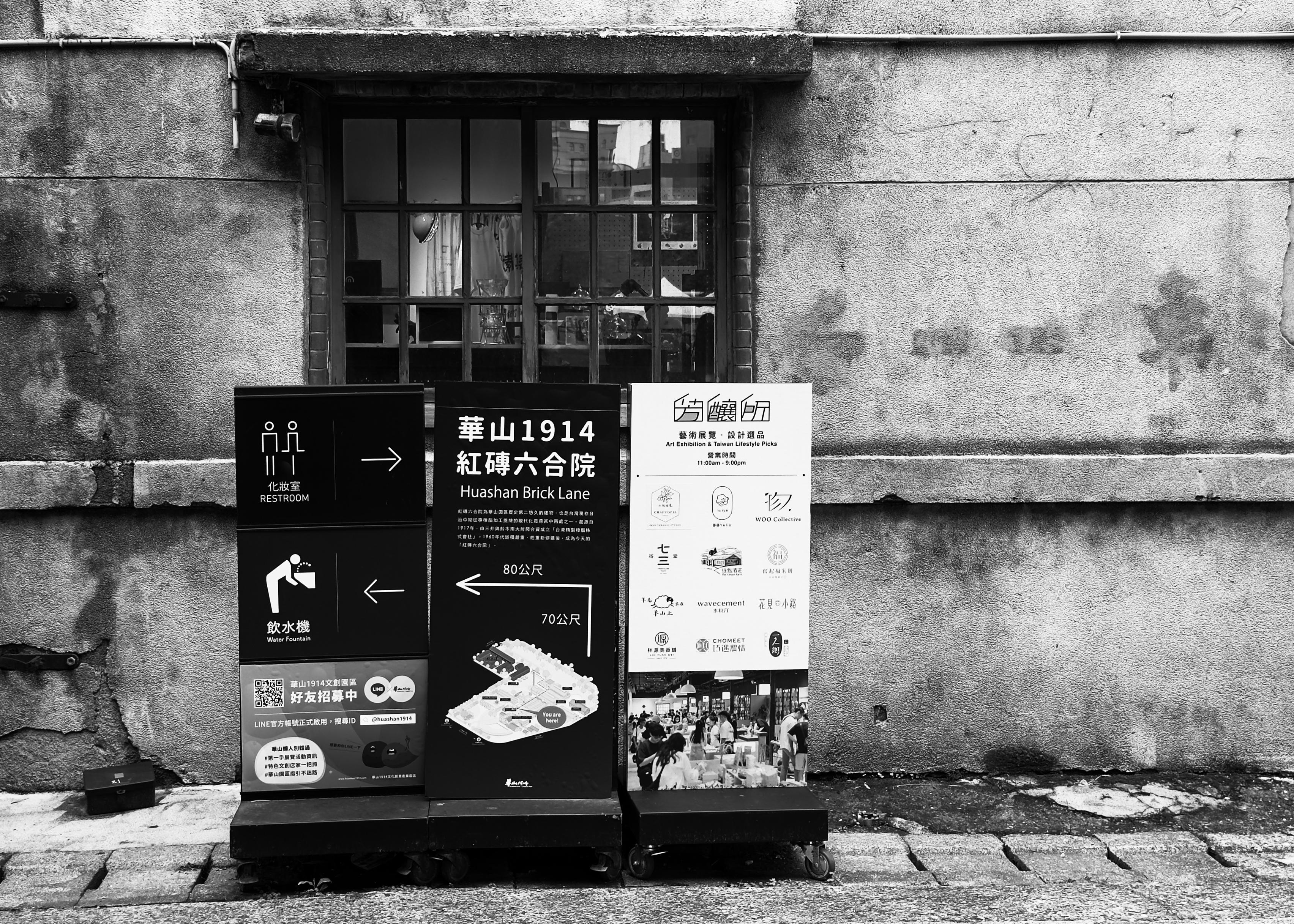 Black and white street scene with informational signs in front of a weathered building wall, including directions and event details.