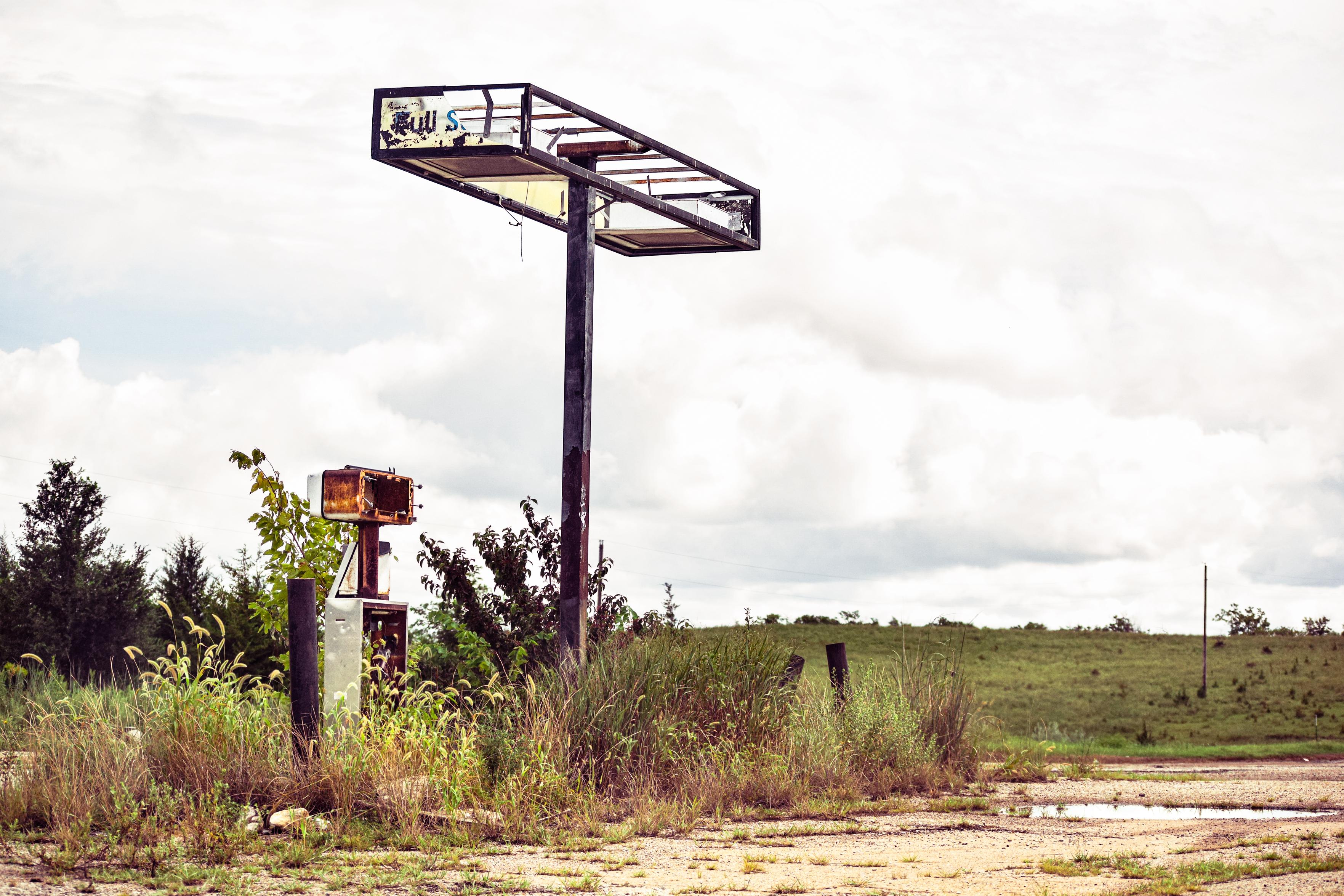 An empty, weathered gas pump frame stands beside an overgrown rural road, with a small sign, open fields, and a quiet, cloudy sky.