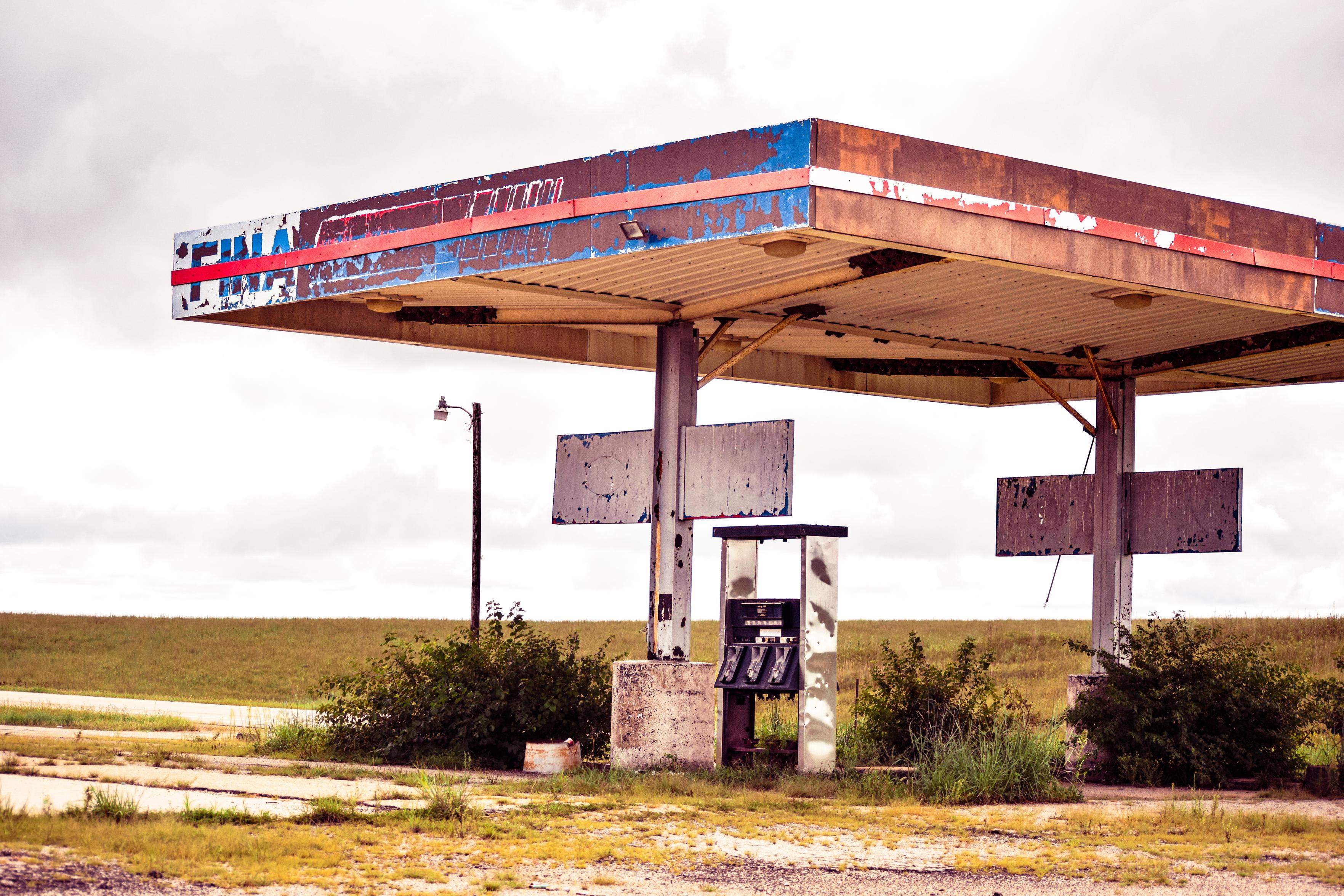 Abandoned roadside gas station with a rusted red-and-blue canopy, broken pumps, and empty signs in a flat field under cloudy skies; desolate mood.