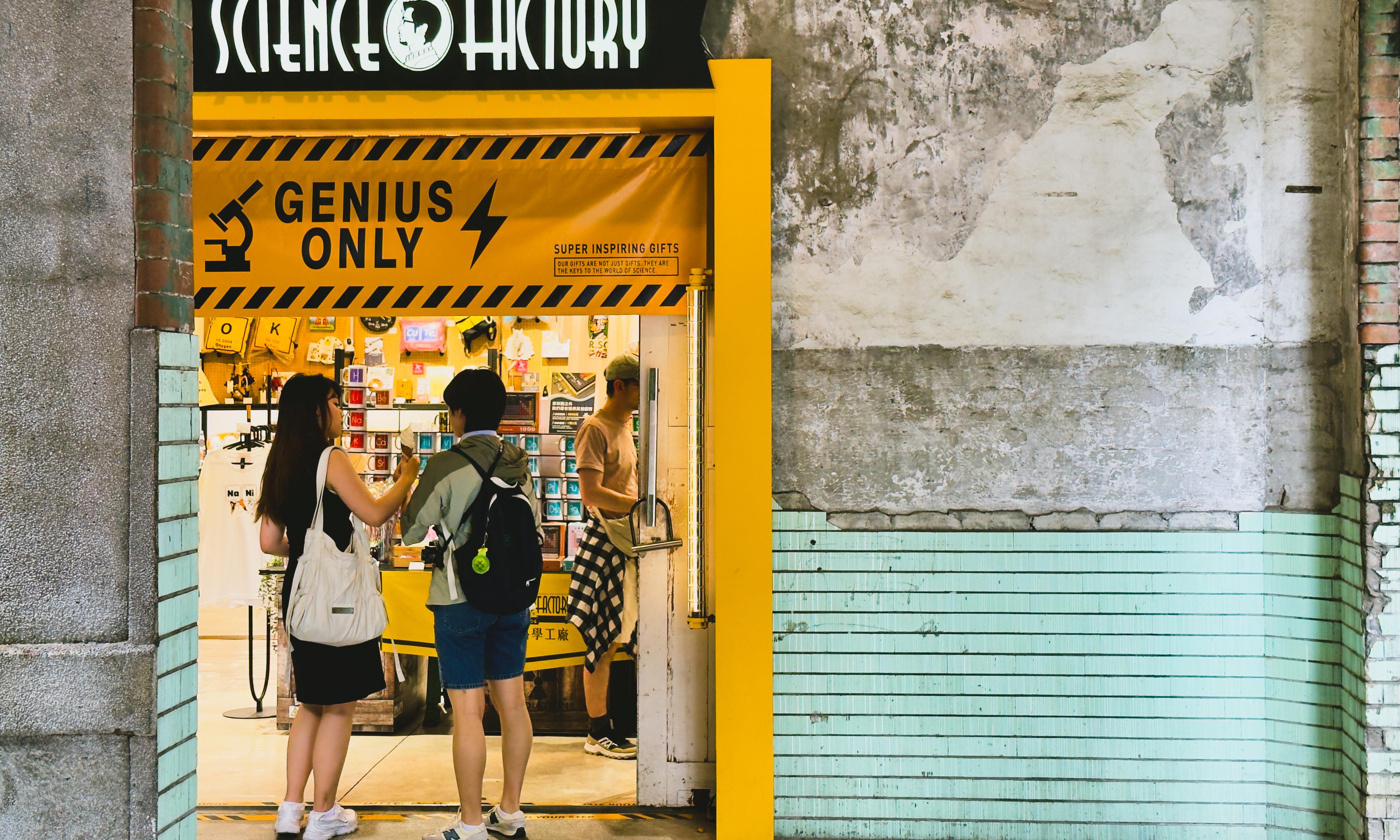Two people with backpacks are shopping at a small store called "Science Factory," which has a bright yellow sign reading "Genius Only" outside.