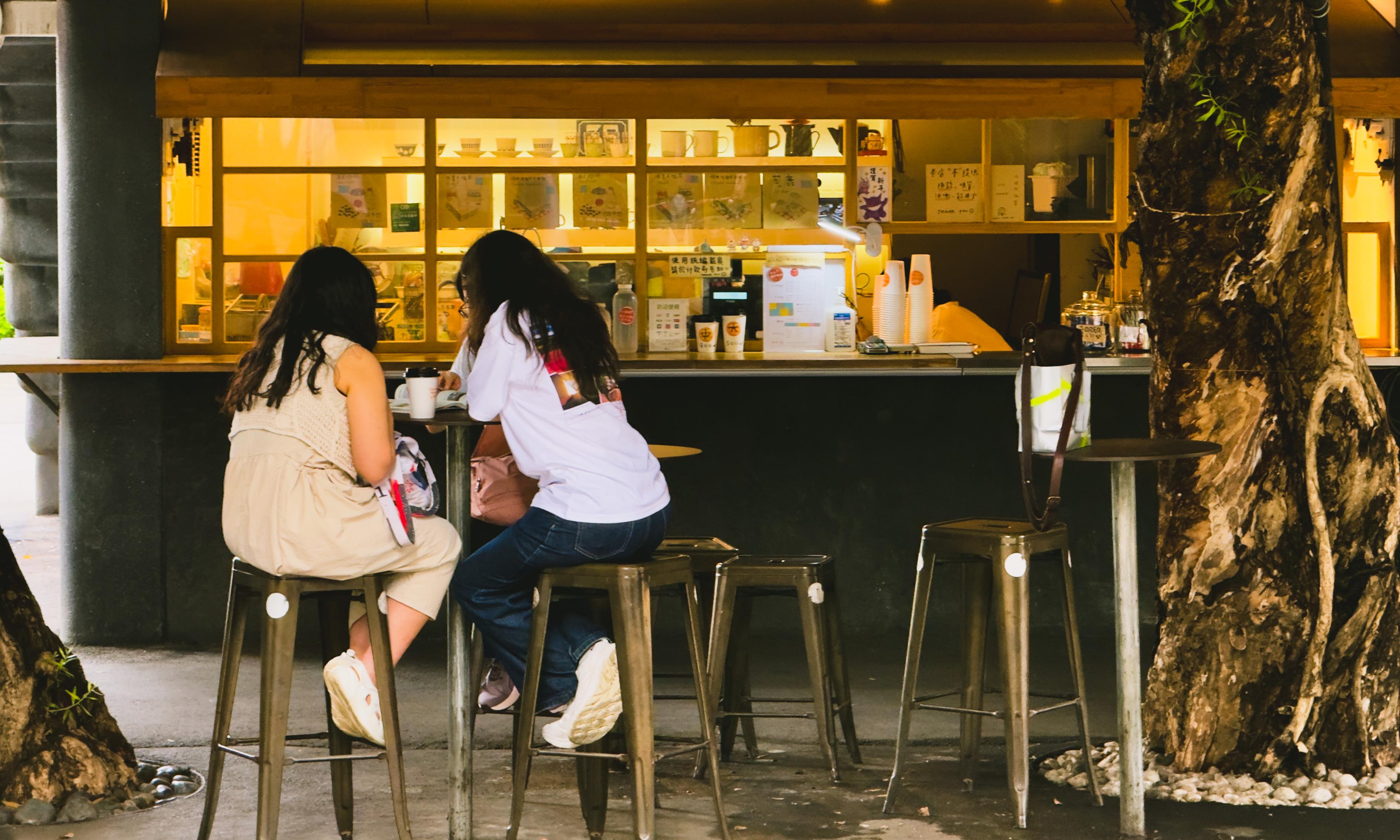 Two women sitting at a bar outside a cafe, engaging in conversation, with trees and illuminated cafe window in the background.